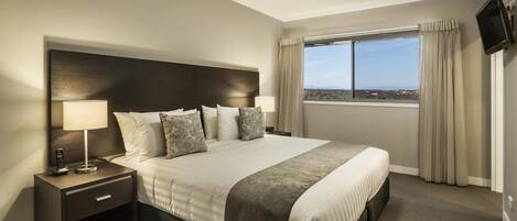 A hotel room with a large bed, two bedside tables with lamps, a TV mounted on the wall, and a window with curtains.