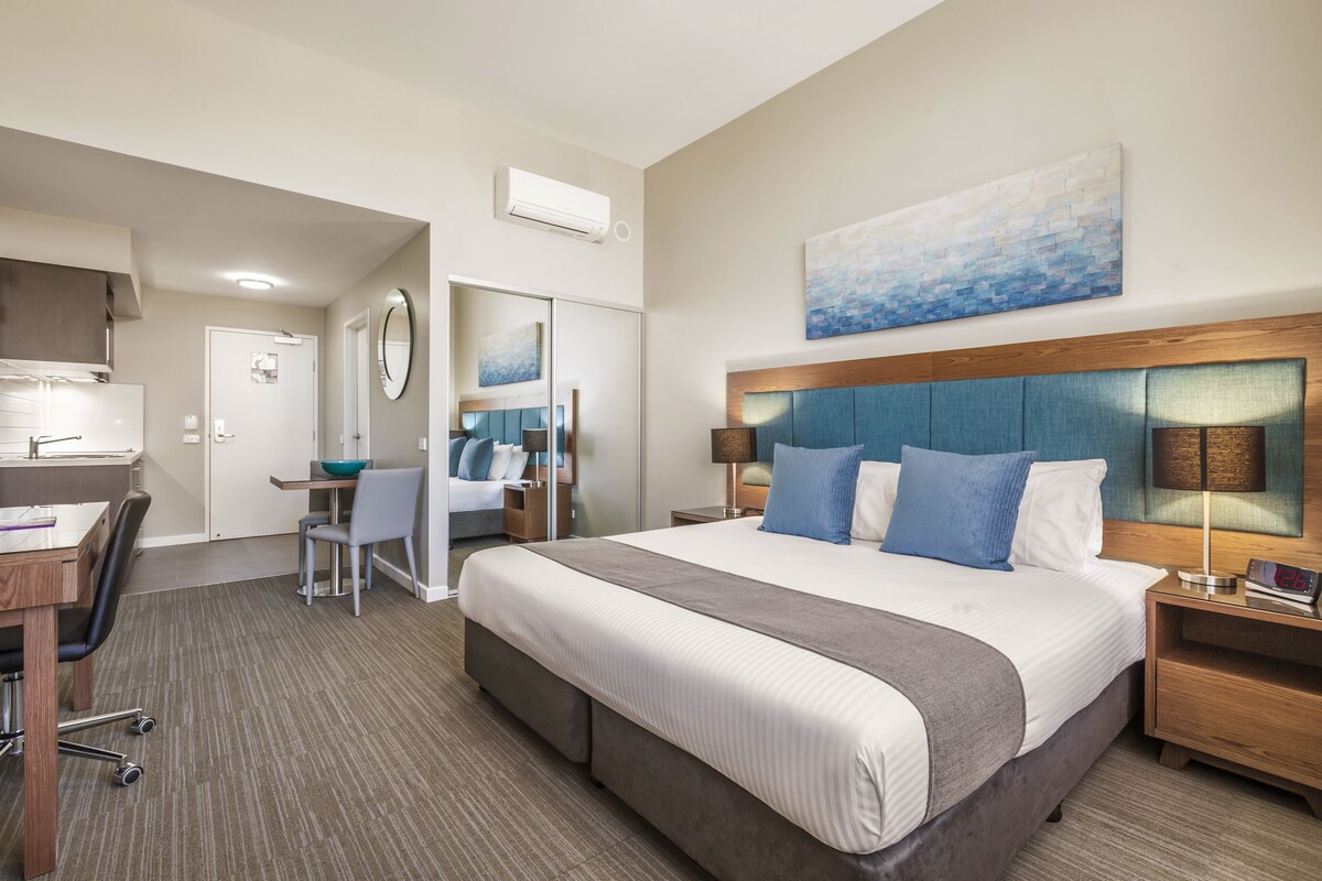 A hotel room with a large bed, a desk, a chair, a mirror, and a painting on the wall.