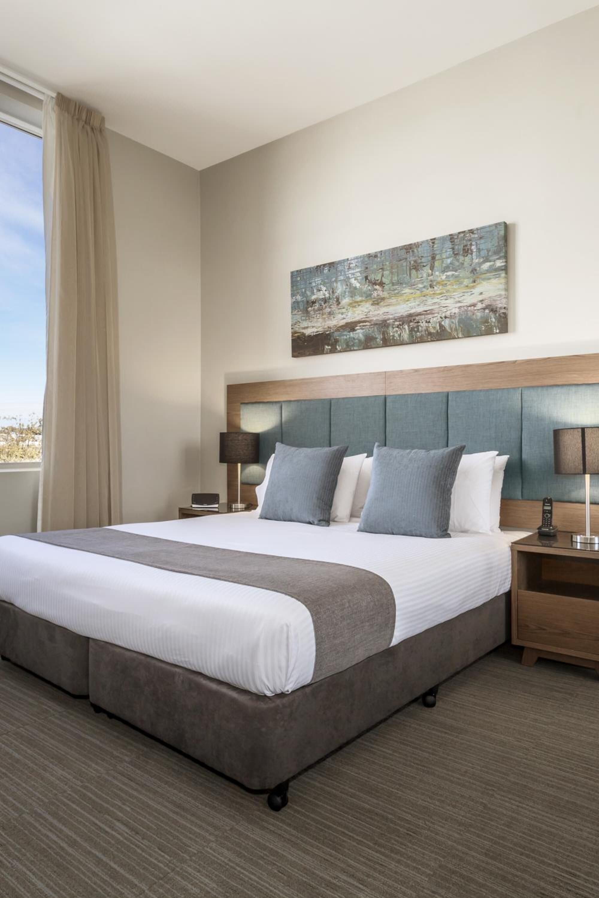 Une chambre d’hôtel avec un grand lit, deux tables de chevet, une tête de lit, une fenêtre avec des rideaux et un tableau au mur.