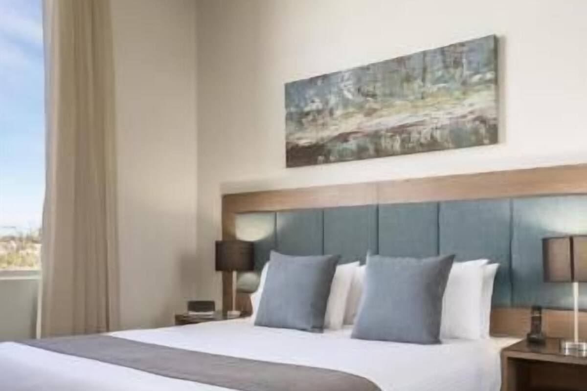 Quarto de hotel com uma cama grande, cabeceira, duas mesas de cabeceira com luminárias, um quadro na parede e uma janela com cortinas.