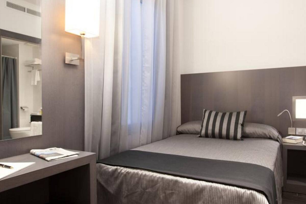 A modern hotel room with a large bed, a desk, a bedside table, a mirror, and a window with curtains.