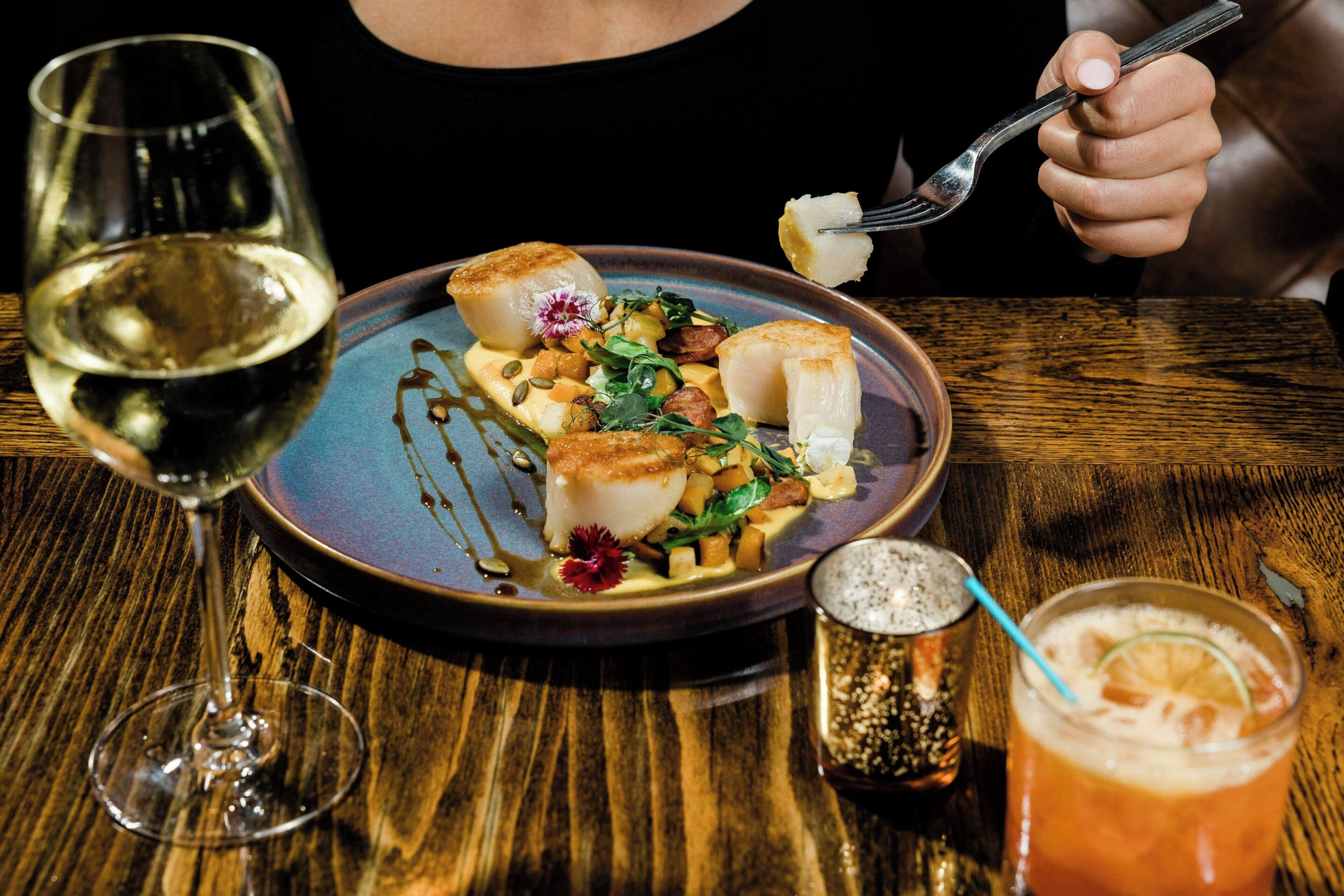 A dining table with a plate of food, a glass of white wine, and a cocktail with a straw.