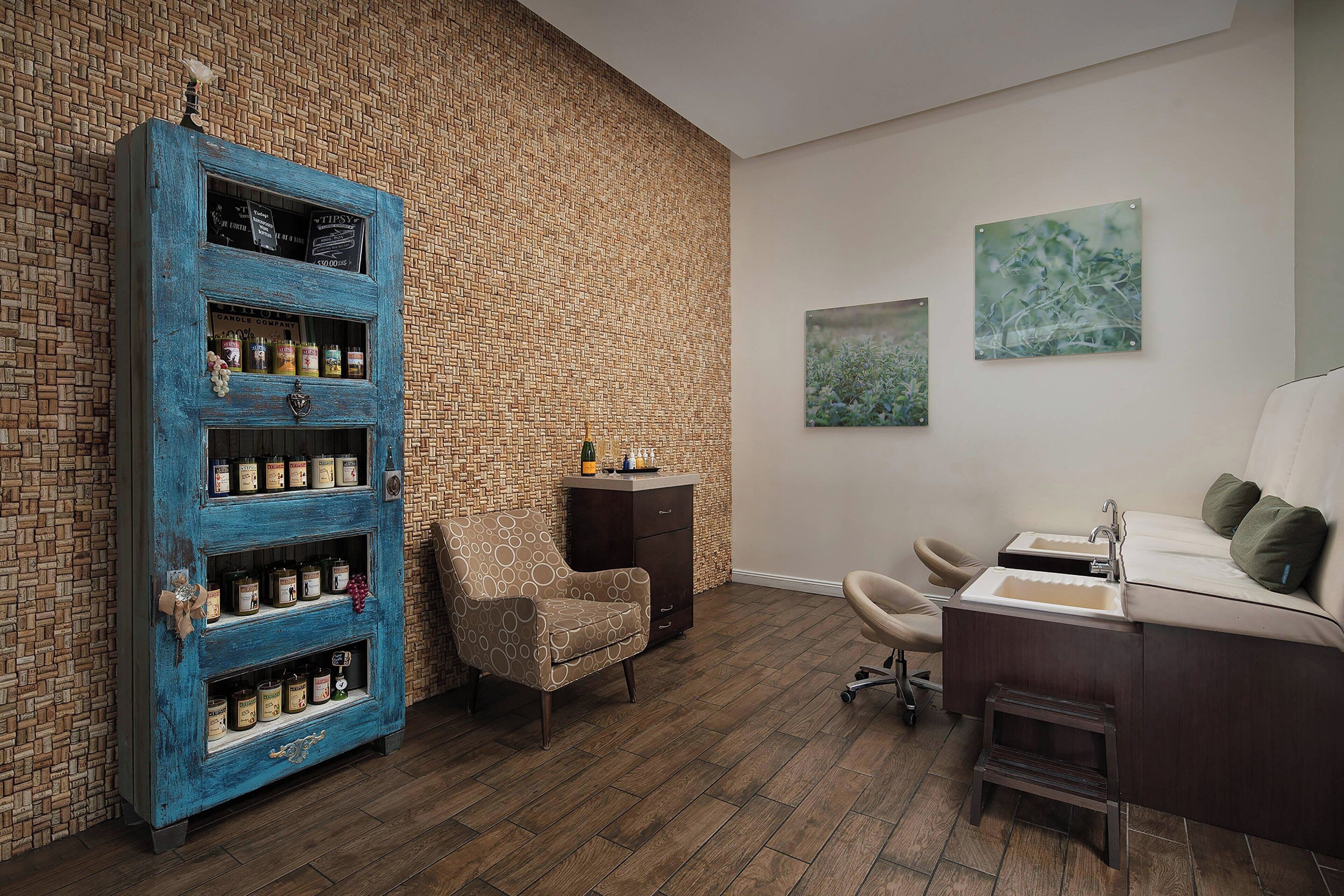A room with a brick-patterned wall, a blue cabinet with multiple shelves, a small armchair, a desk with a sink, and two framed artworks.
