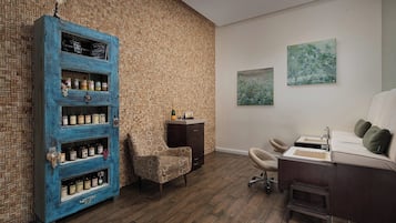 A room with a brick-patterned wall, a blue cabinet with multiple shelves, a small armchair, a desk with a sink, and two framed artworks.