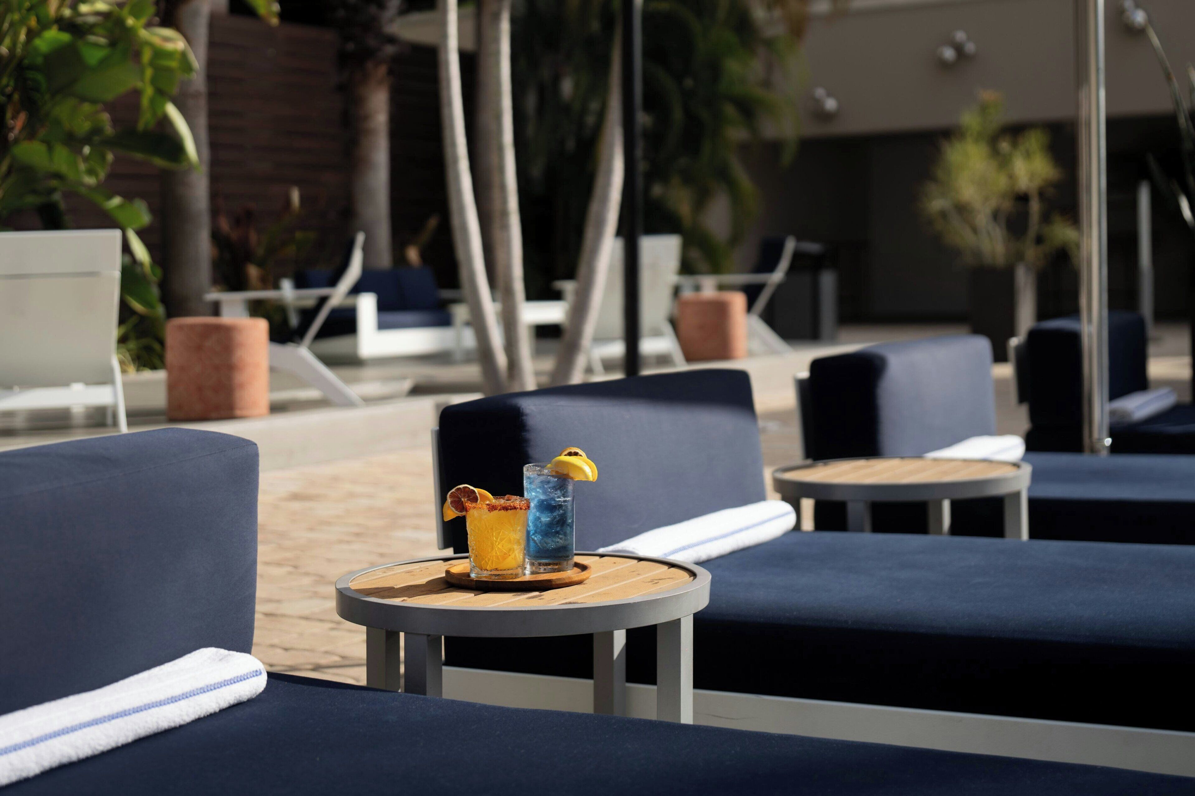A poolside lounge area with blue cushioned chairs, a small round table with a drink, and a wooden chair.