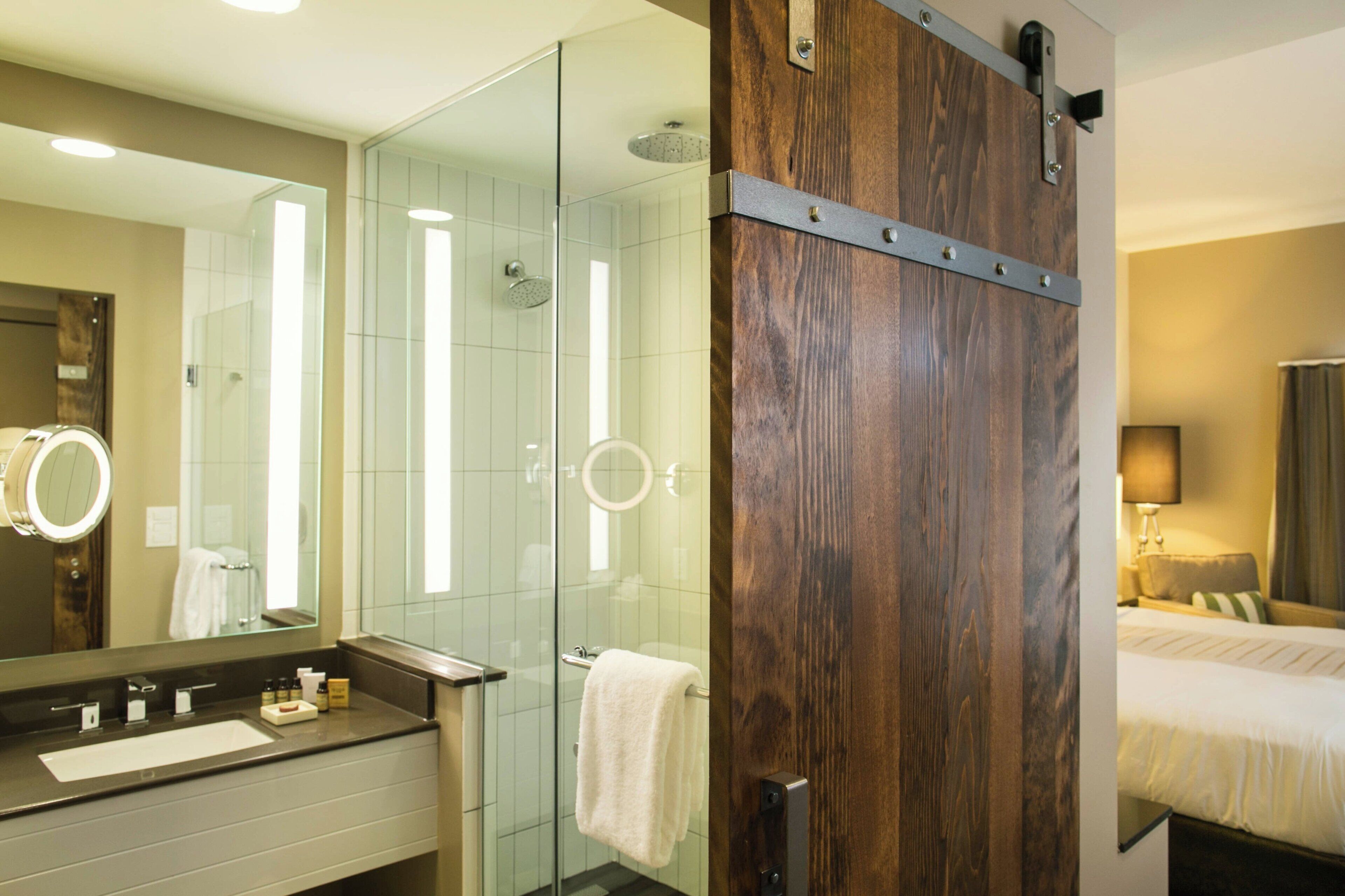 A hotel bathroom with a glass shower, a wooden door, a sink, and a mirror.