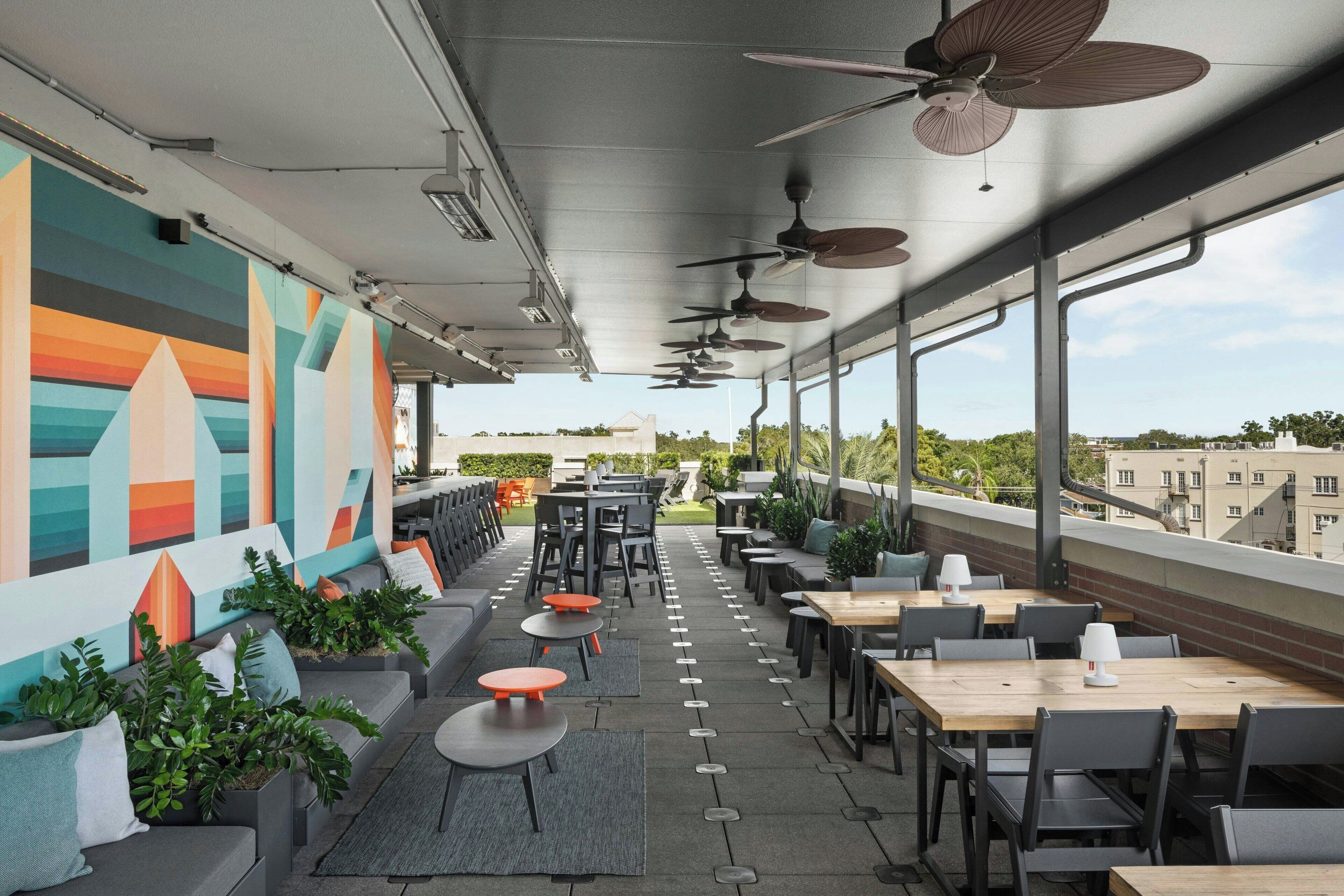An outdoor seating area with tables and chairs, ceiling fans, and a colorful mural on the wall.