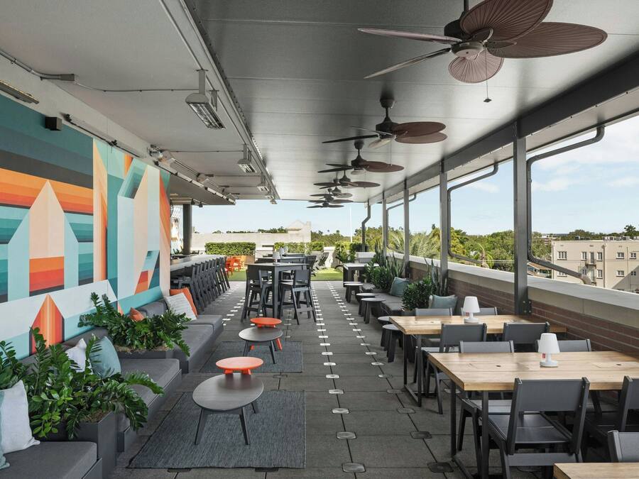 An outdoor seating area with tables and chairs, ceiling fans, and a colorful mural on the wall.