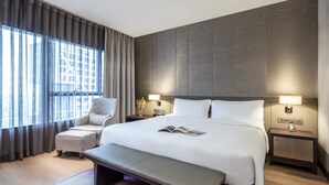 A modern hotel room with a large bed, a bedside table with a lamp, a chair, and a bench.