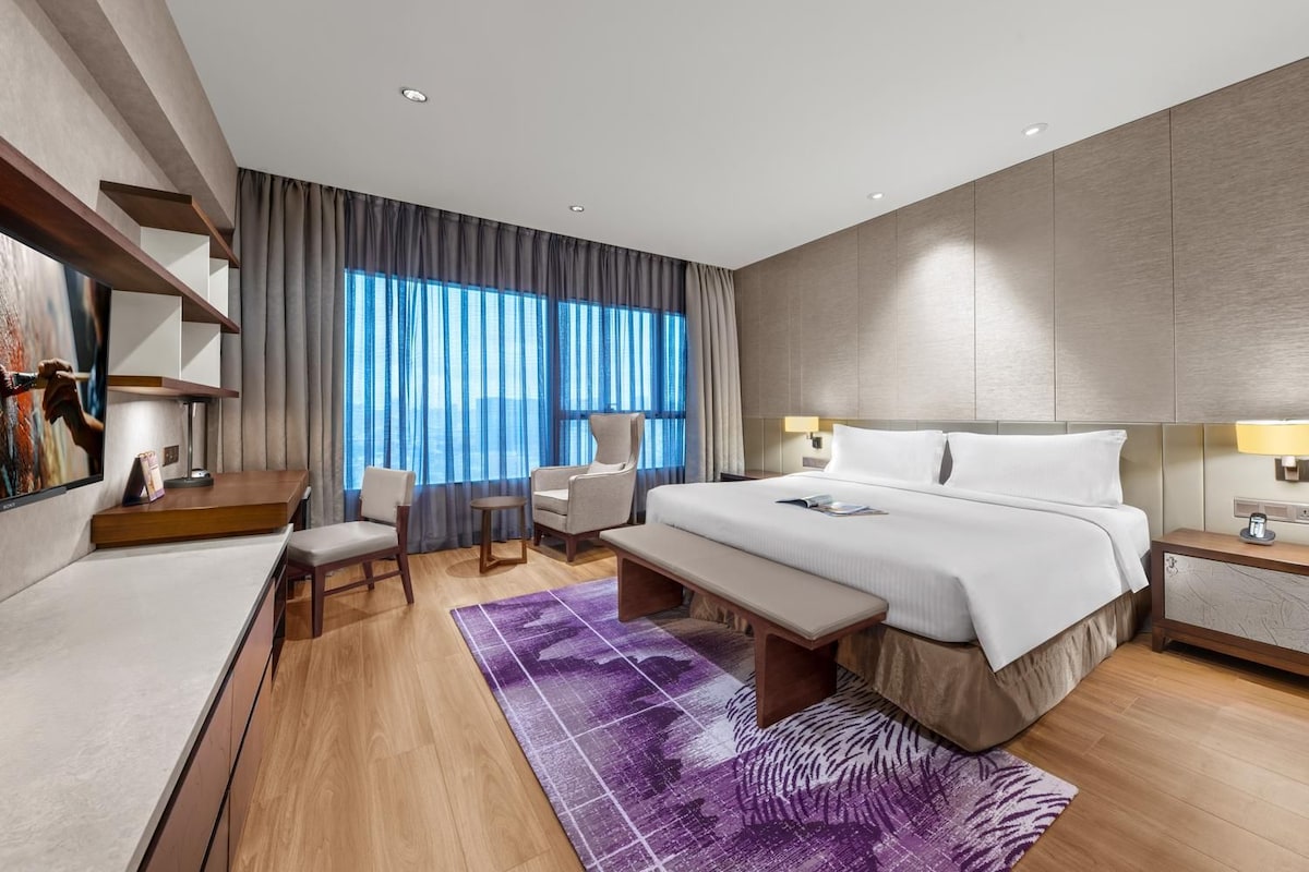 Una habitación de hotel moderna con una cama grande, un escritorio, dos sillas, un banco y vistas de la ciudad a través de amplios ventanales.