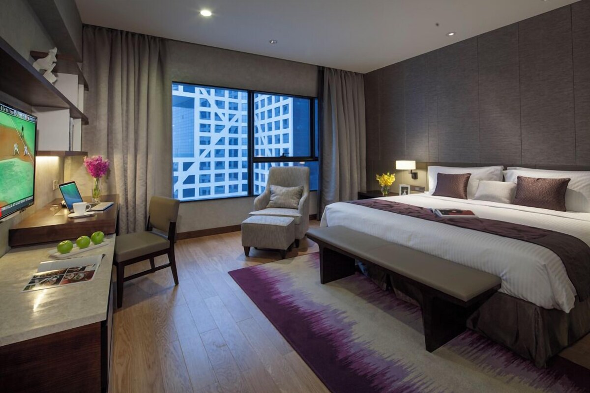 Una habitación de hotel moderna con una cama grande, un escritorio con una computadora portátil, una silla y vistas a un paisaje urbano a través de ventanas amplias.