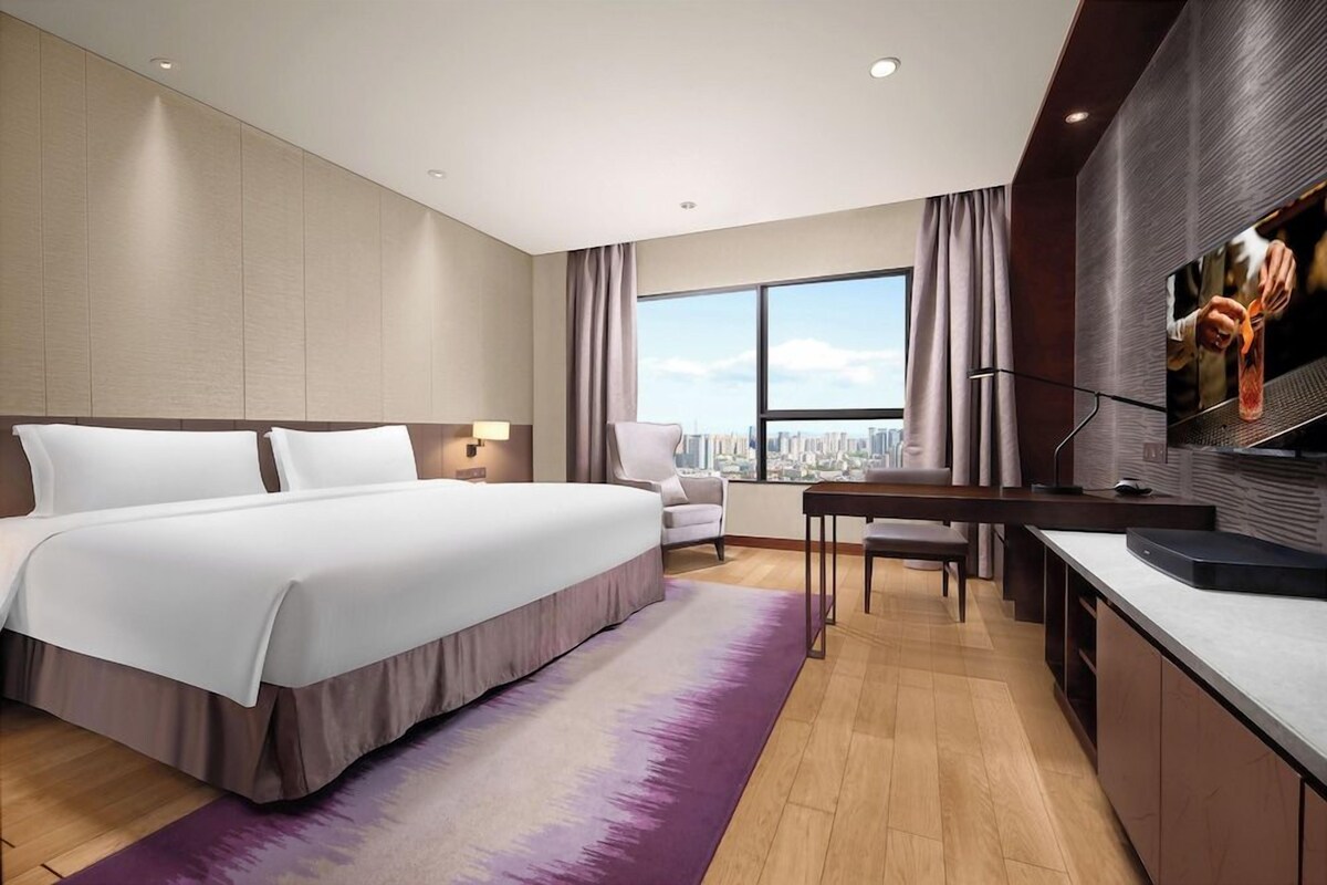 Habitación de hotel moderna con una cama grande, un escritorio, una silla, un televisor y vistas a la ciudad.
