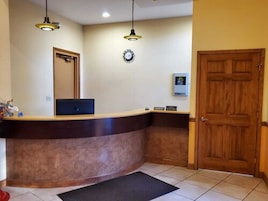 Zona de recepción con un mostrador de madera, dos lámparas colgantes, un reloj, una puerta y un pequeño letrero.