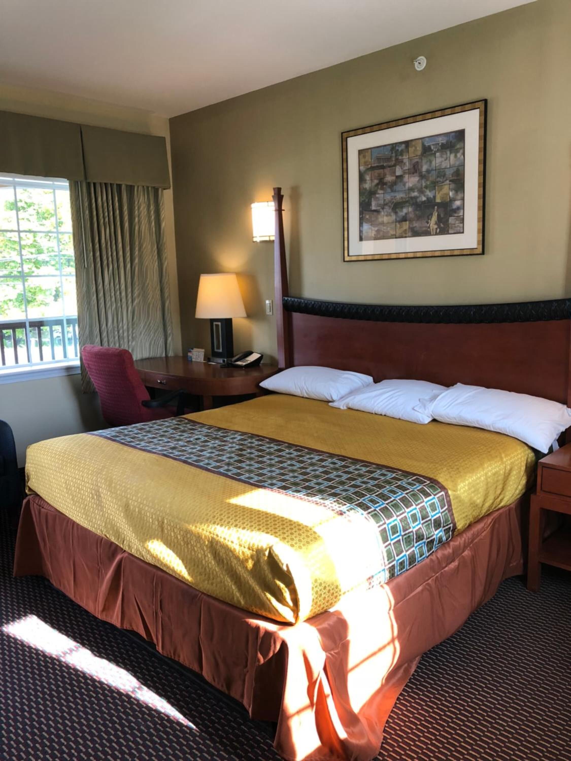 A hotel room with a bed, a desk with a lamp, a chair, and a window with curtains.