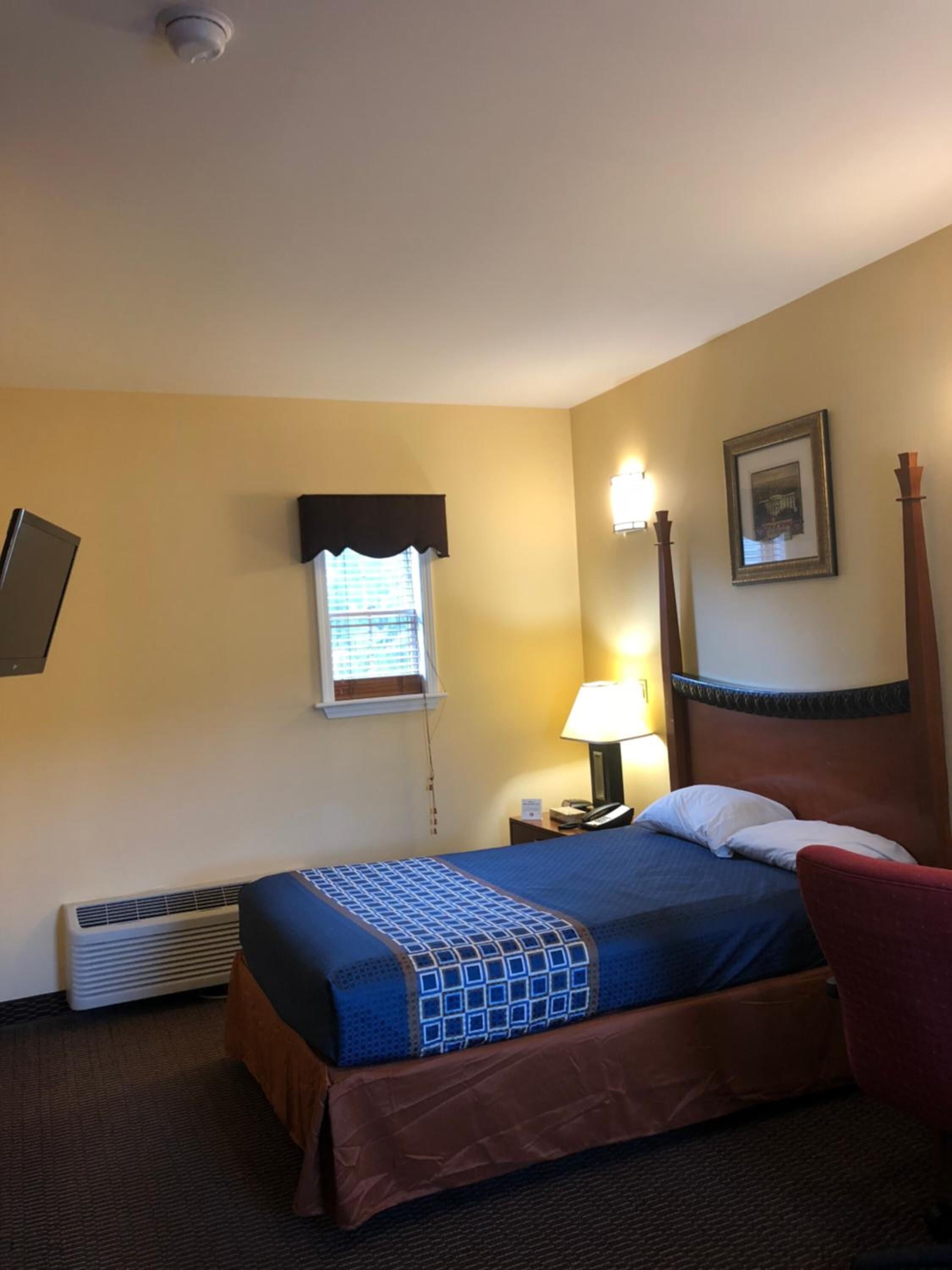 A hotel room with a bed, a bedside table with a lamp, a window with blinds, a wall-mounted TV, and a framed picture.