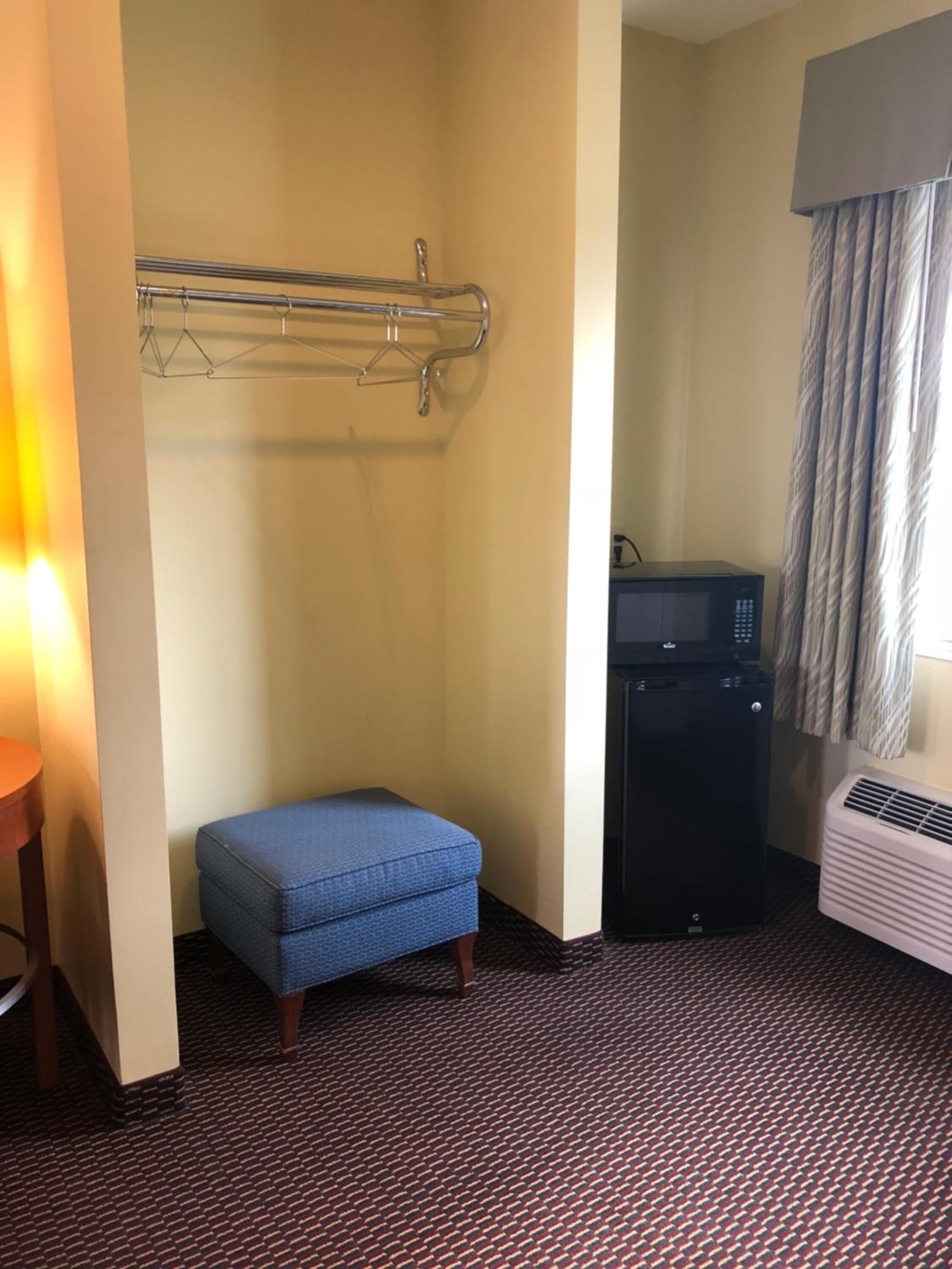 Quarto de hotel com um pequeno banco azul, micro-ondas, aquecedor e suporte de toalhas fixado na parede.