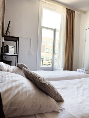 A hotel room with two beds, a window with curtains, a shelf with a kettle, and a view of buildings outside. - Hotel Amsterdam Inn - Digital Access (Amsterdam)