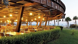 An outdoor restaurant with wooden tables and chairs, a curved roof structure, and a view of the sea. - Mukarnas Spa & Resort (Alanya)