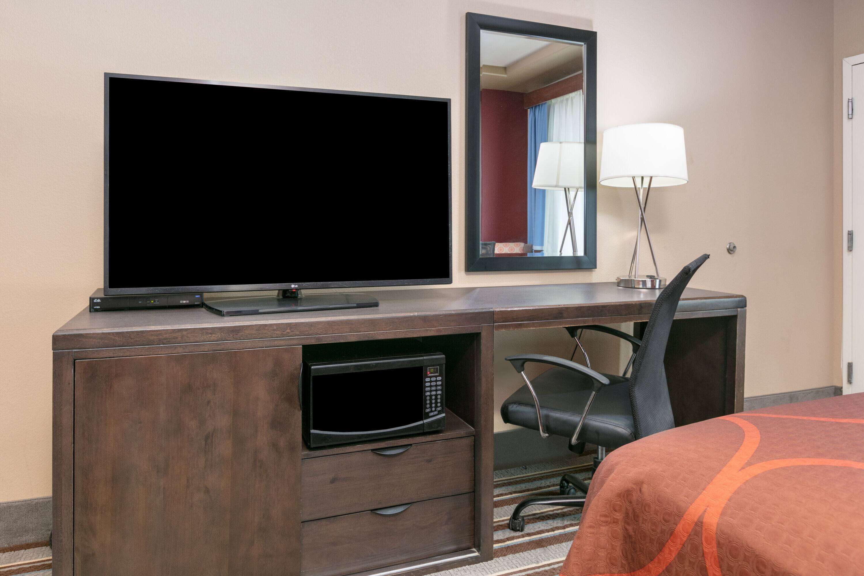 A hotel room with a flat-screen TV, a microwave, a desk with a chair, a bed with an orange blanket, and a mirror.