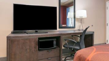 A hotel room with a flat-screen TV, a microwave, a desk with a chair, a bed with an orange blanket, and a mirror.