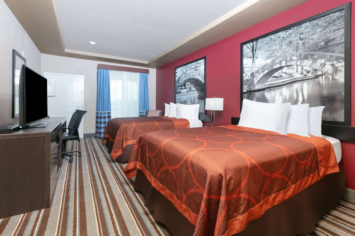 A hotel room with two beds, a desk, a TV, and a large window with curtains.