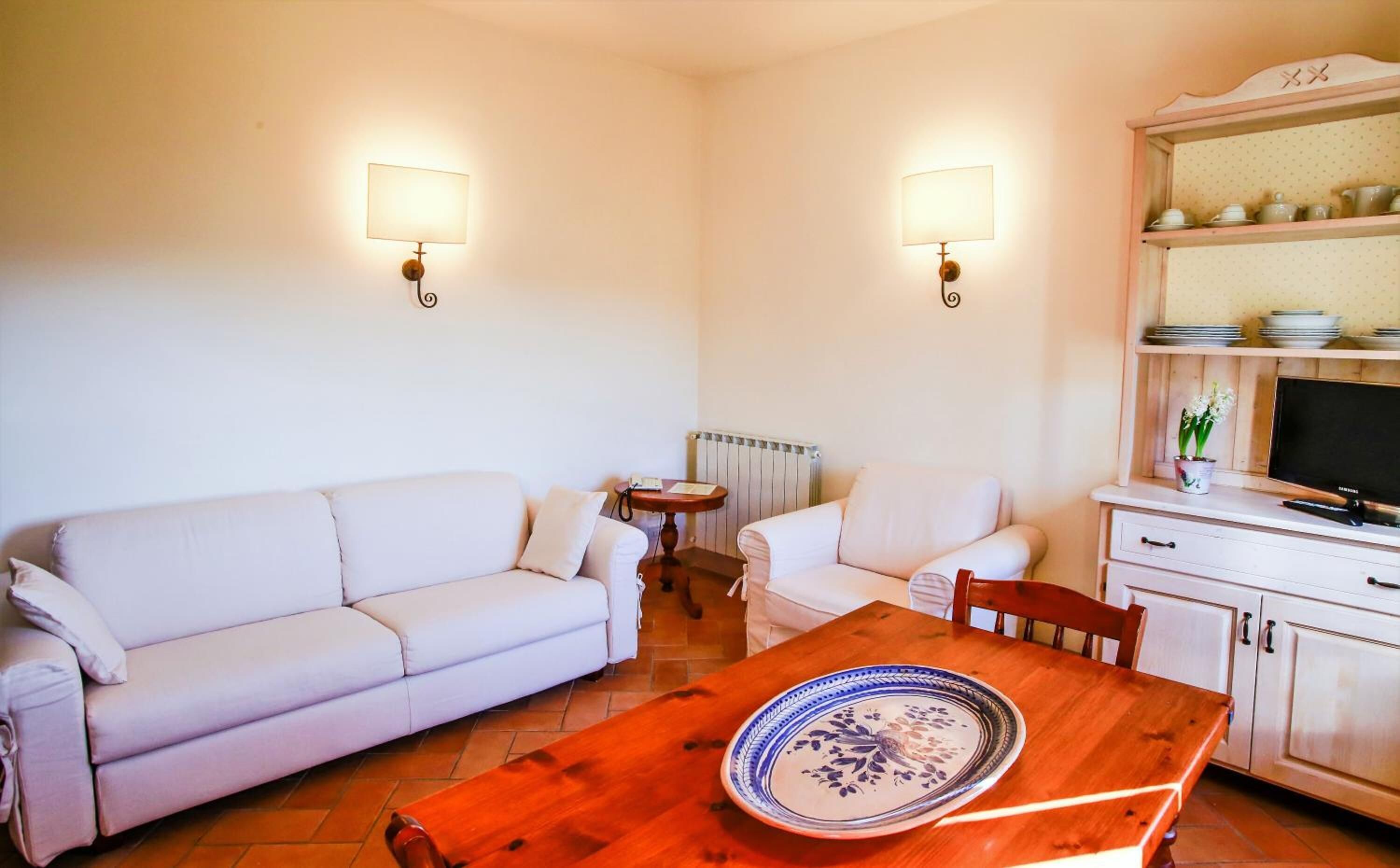 A living room with a sofa, two armchairs, a wooden table with a decorative plate, a small side table, a television, and a kitchen area with a shelf and a radiator.