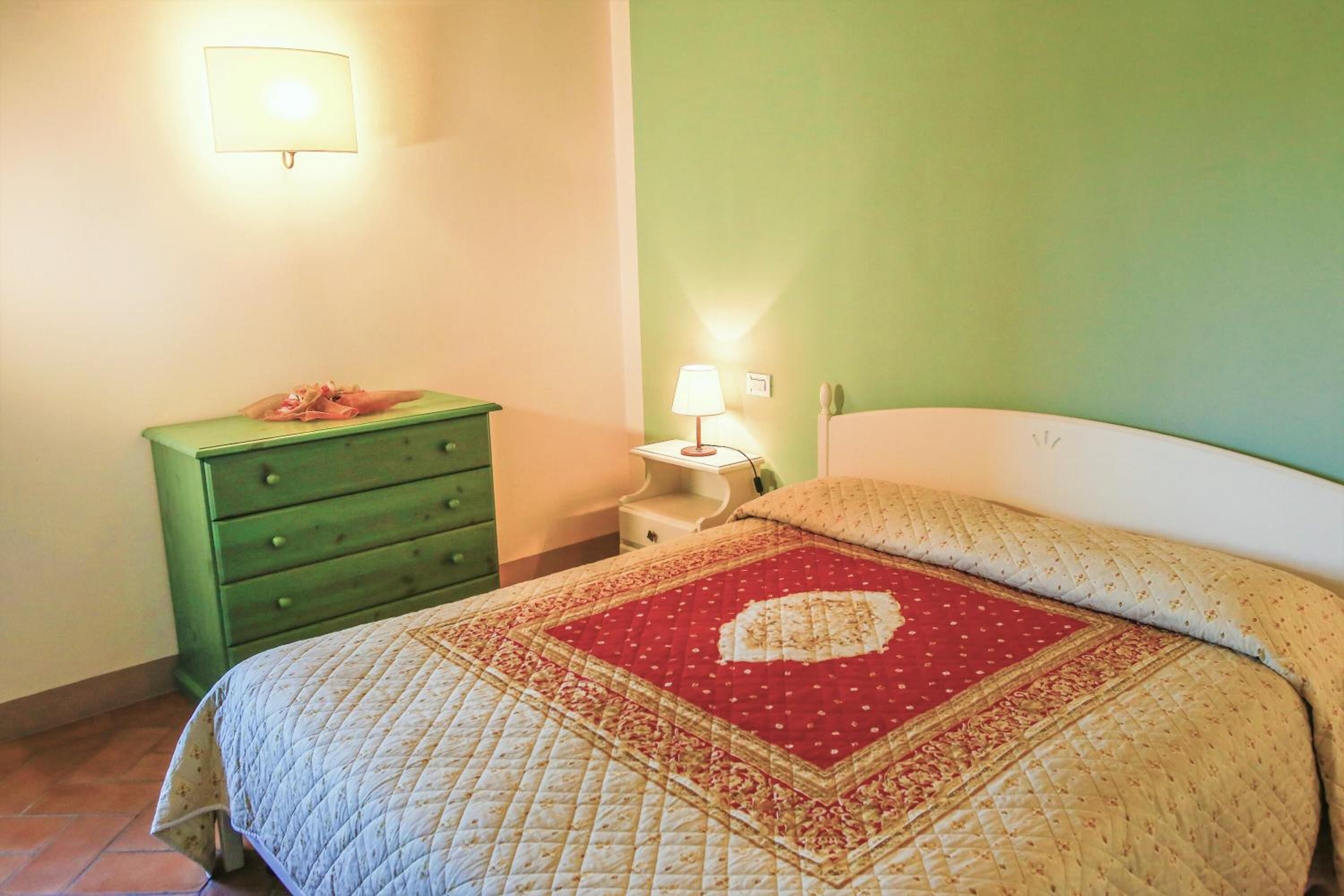 A bedroom with a bed, a green dresser, a nightstand with a lamp, and a wall-mounted light.