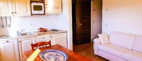 Une cuisine compacte avec des meubles blancs, une table à manger en bois et un canapé blanc dans l’espace salon.