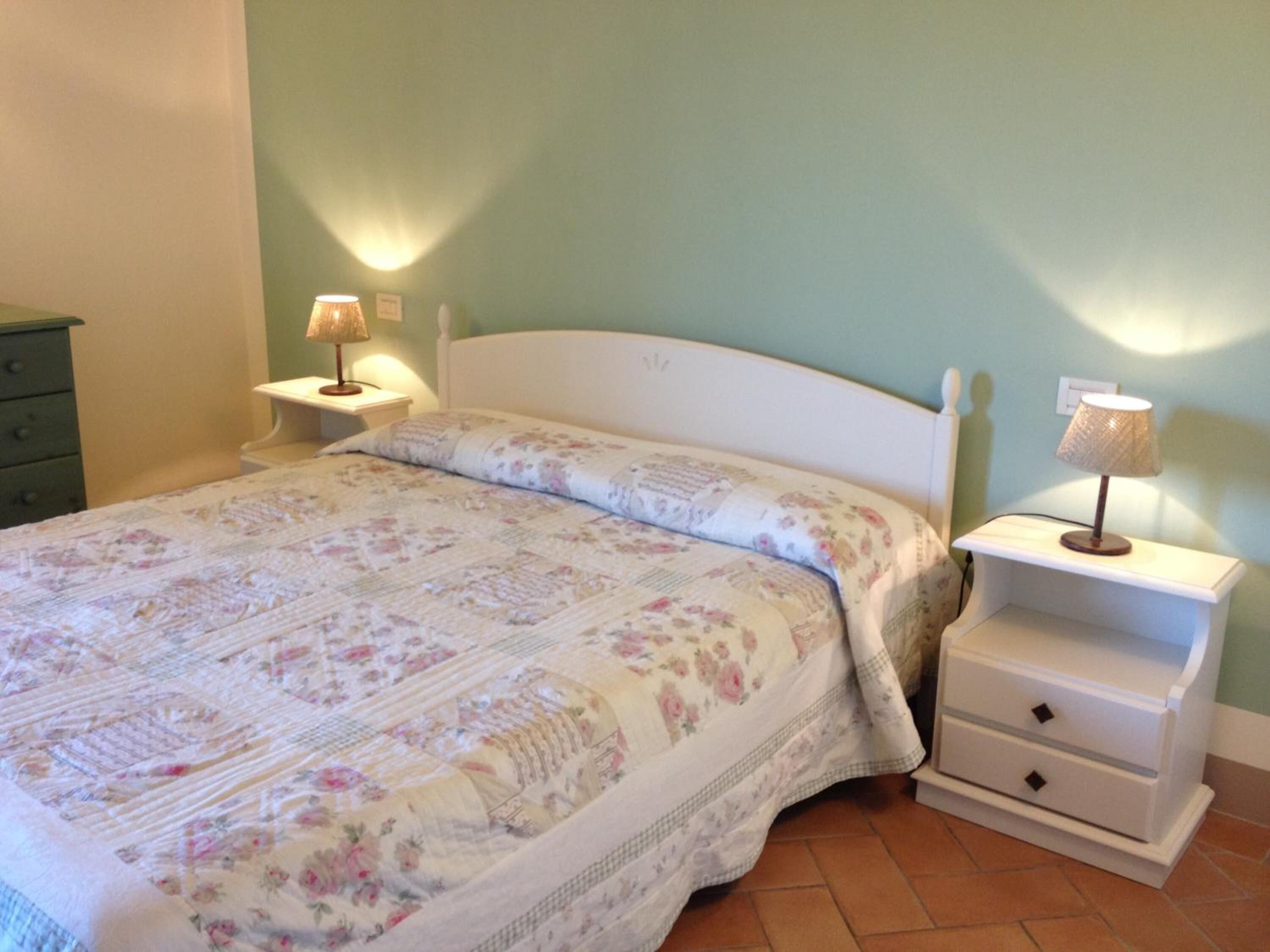Un letto rifatto con biancheria floreale, due comodini con lampade e una cassettiera verde.