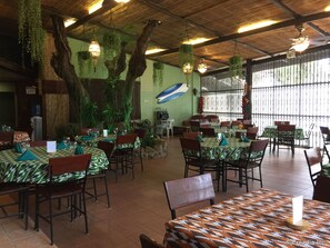 Un restaurante con mesas cubiertas por manteles estampados, plantas colgantes y techo de paja.