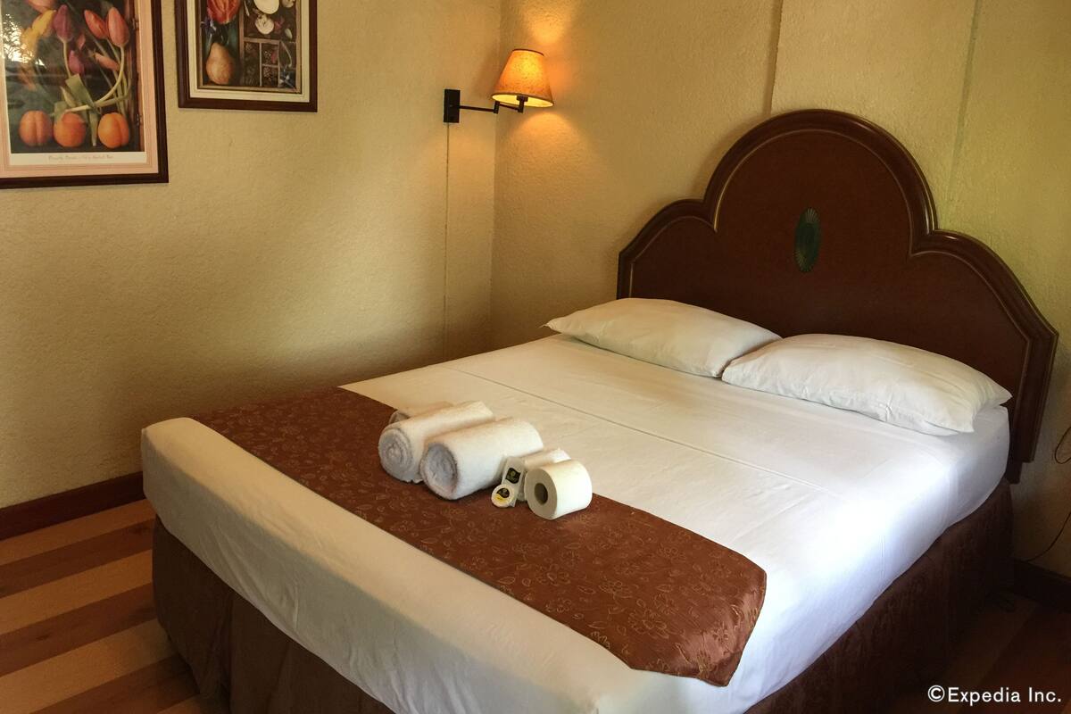Ein ordentlich bezogenes Bett mit weißer Bettwäsche und einem braunen Zierläufer, eine Wandlampe und zwei gerahmte Pflanzenbilder an der Wand.