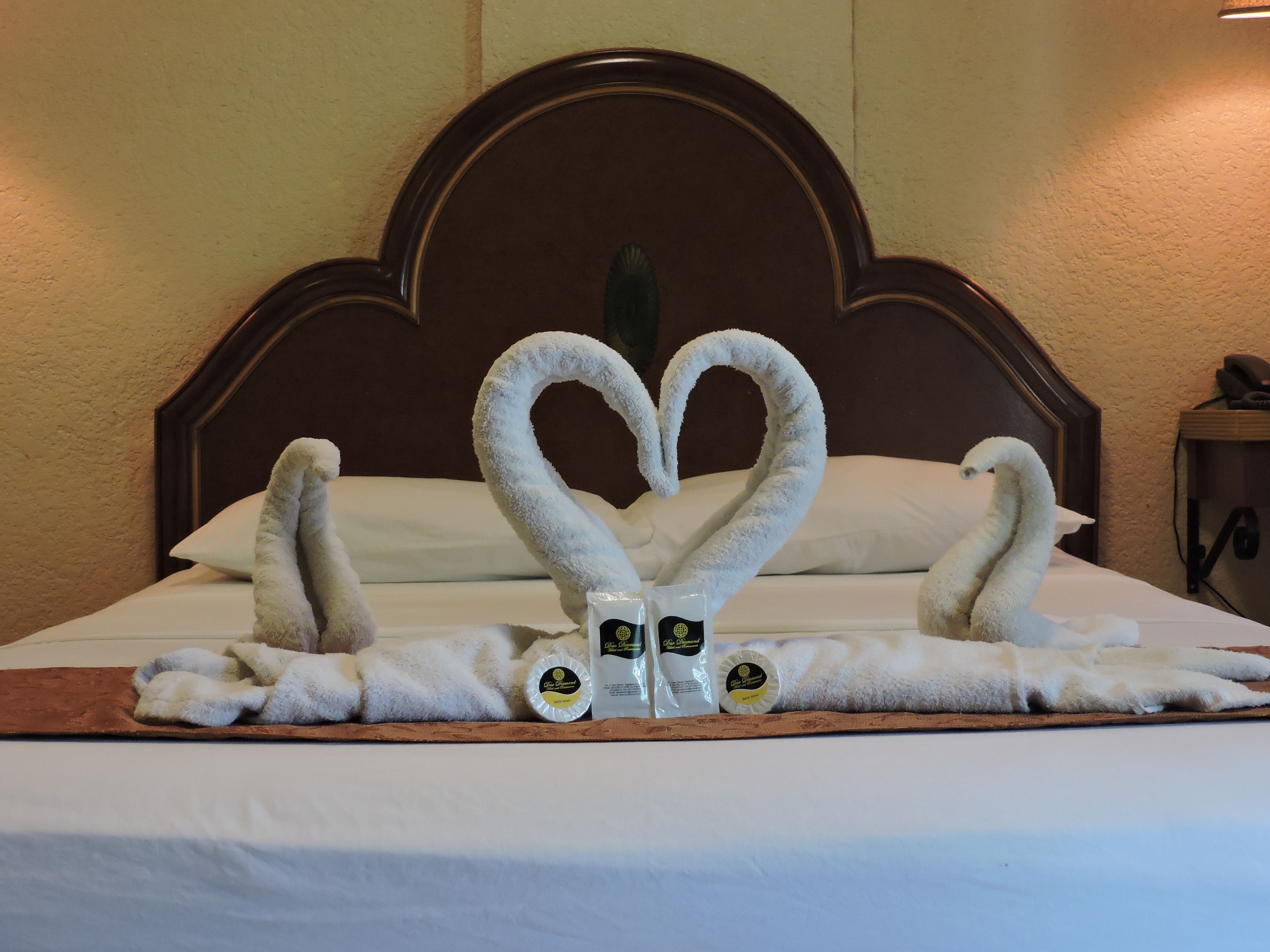 Une chambre d’hôtel avec un lit agrémenté d’une décoration en forme de cœur réalisée avec des serviettes, ainsi que deux bouteilles de boissons proposées dans le minibar.