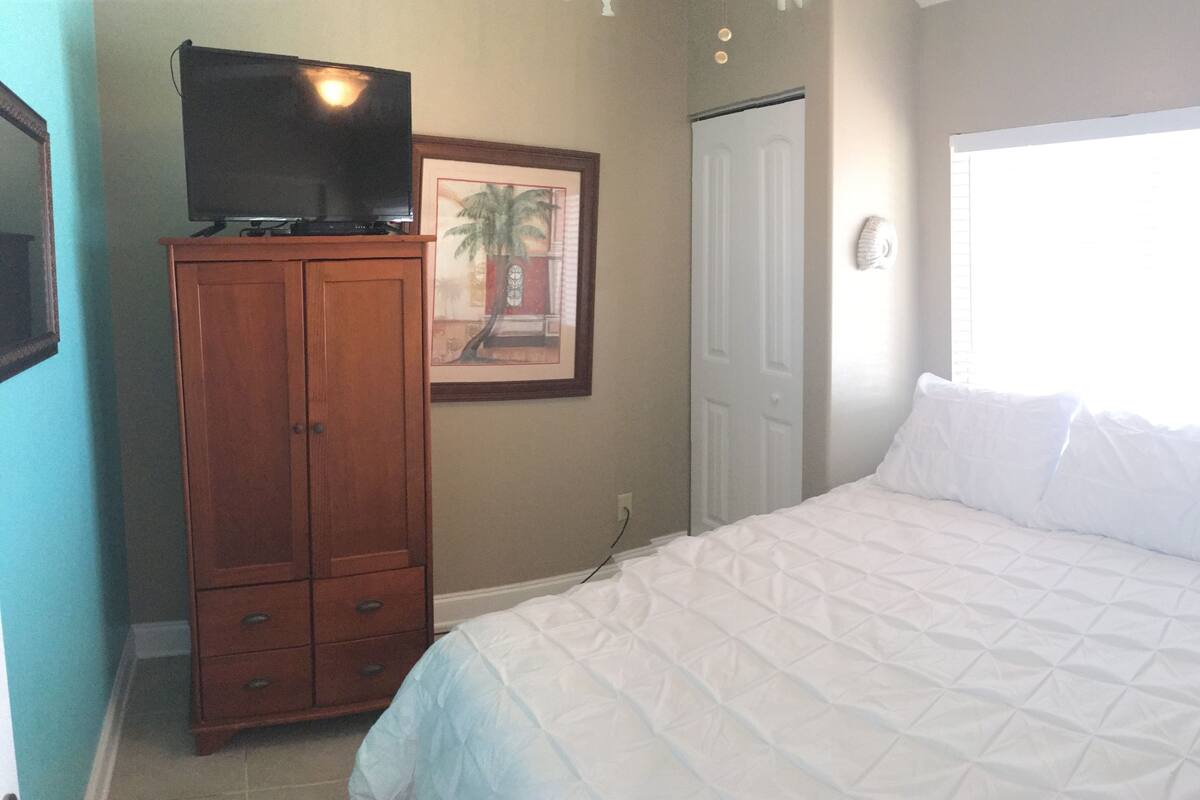 Une chambre à coucher avec un lit, une armoire en bois, un téléviseur posé sur l’armoire, un tableau encadré accroché au mur et une porte.