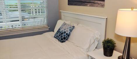 A neatly made bed with white bedding and a blue patterned pillow, a bedside lamp, a small potted plant, and a painting of a stormy sea on the wall.