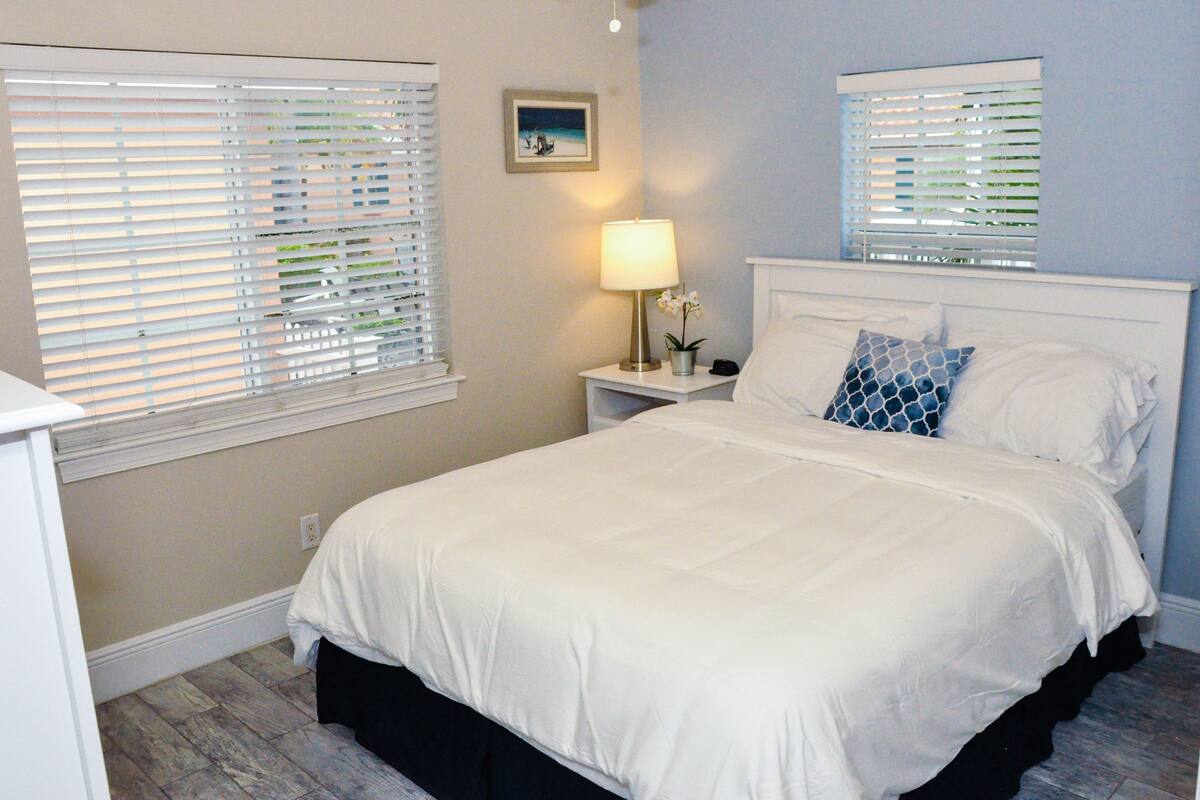 Une chambre à coucher comprenant un lit, deux tables de chevet, une commode, une lampe et deux fenêtres équipées de stores.