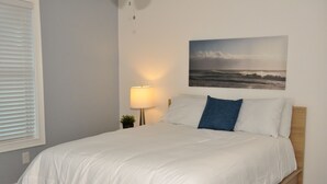 A neatly made bed with white bedding and a blue pillow, a bedside lamp, and a framed picture of the ocean on the wall.