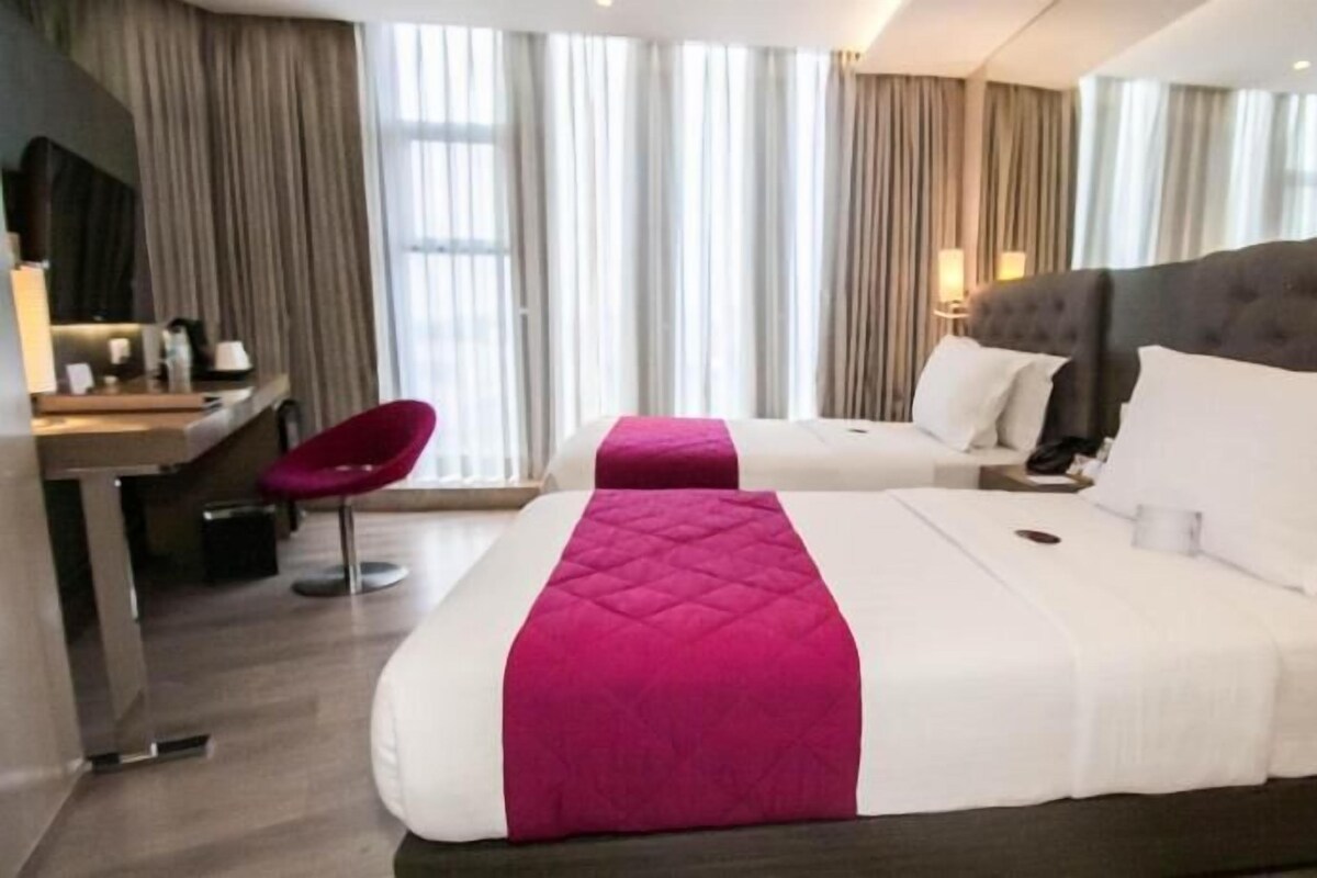 A hotel room with two beds, a desk, a chair, and a large window with curtains.
