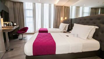 Habitación de hotel con dos camas, un escritorio, una silla y ventanas grandes con cortinas.