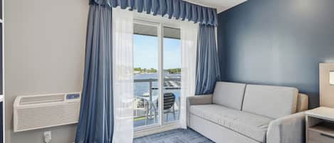 A room with a sofa, a small table, a window with curtains, and a view of a marina.