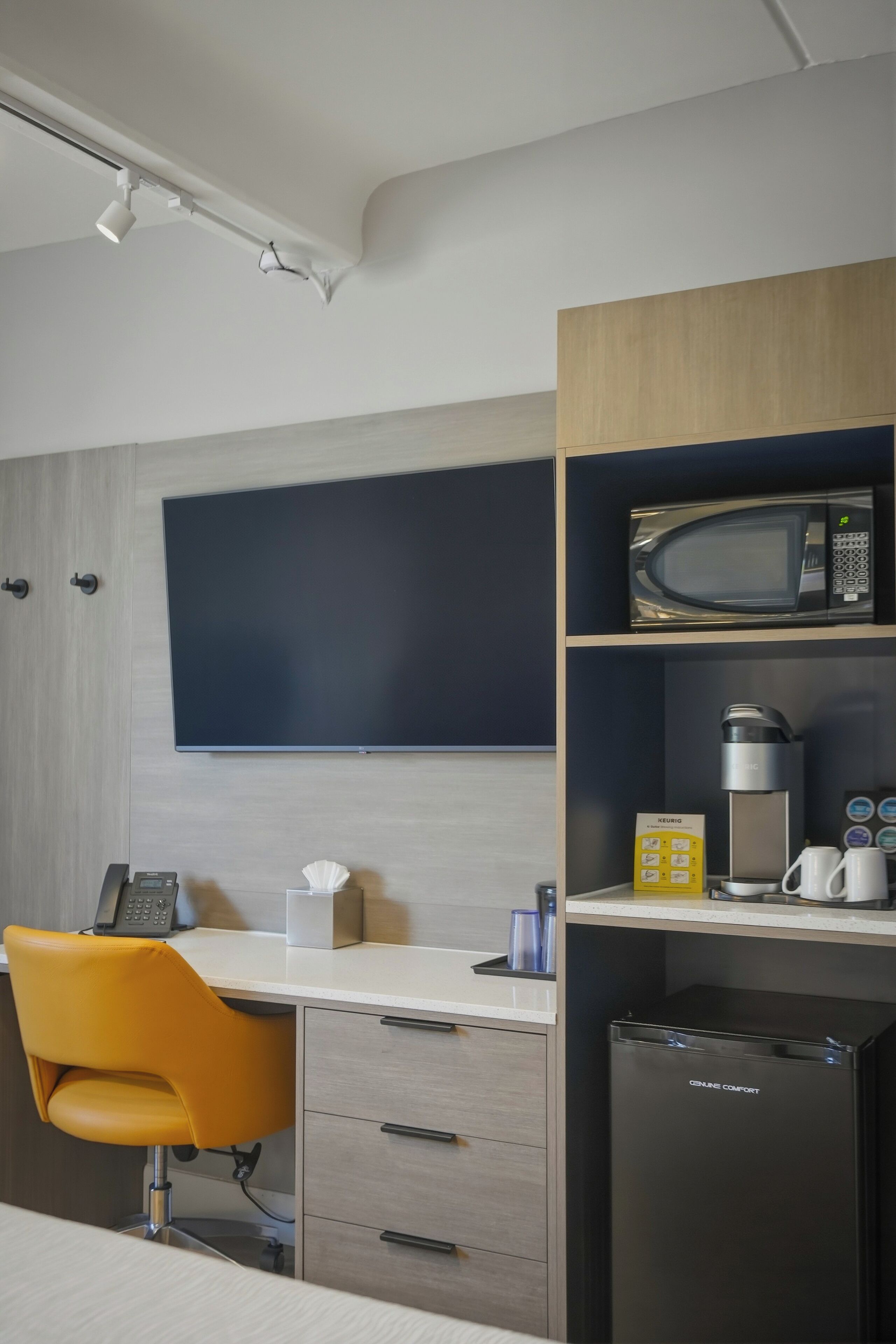 Una habitación de hotel moderna con televisión de pantalla plana, microondas, cafetera, refrigerador pequeño y una silla amarilla.