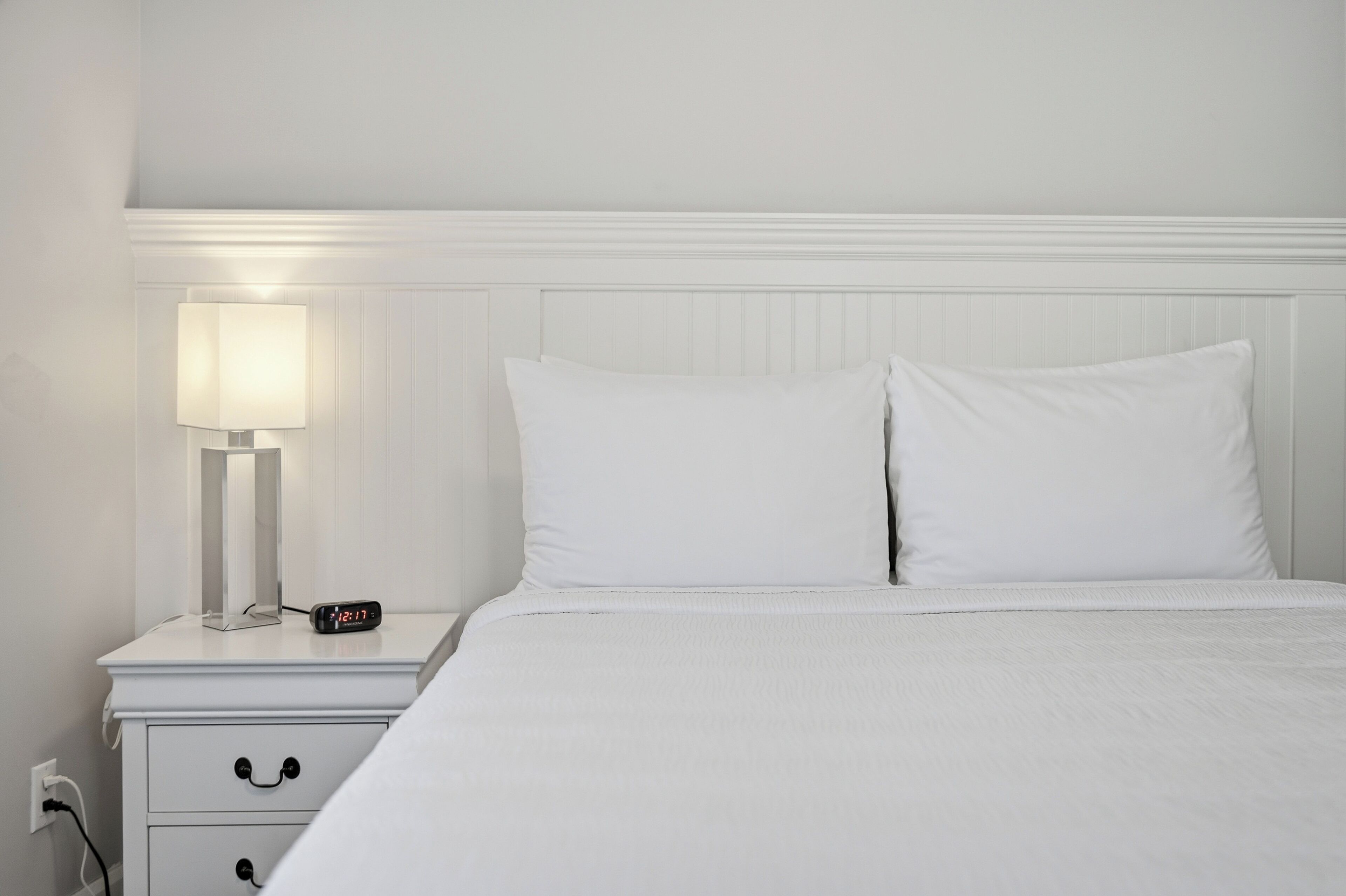 Una cama bien hecha con ropa blanca, una mesita de noche con lámpara y despertador, y una pared blanca con cabecera sencilla.