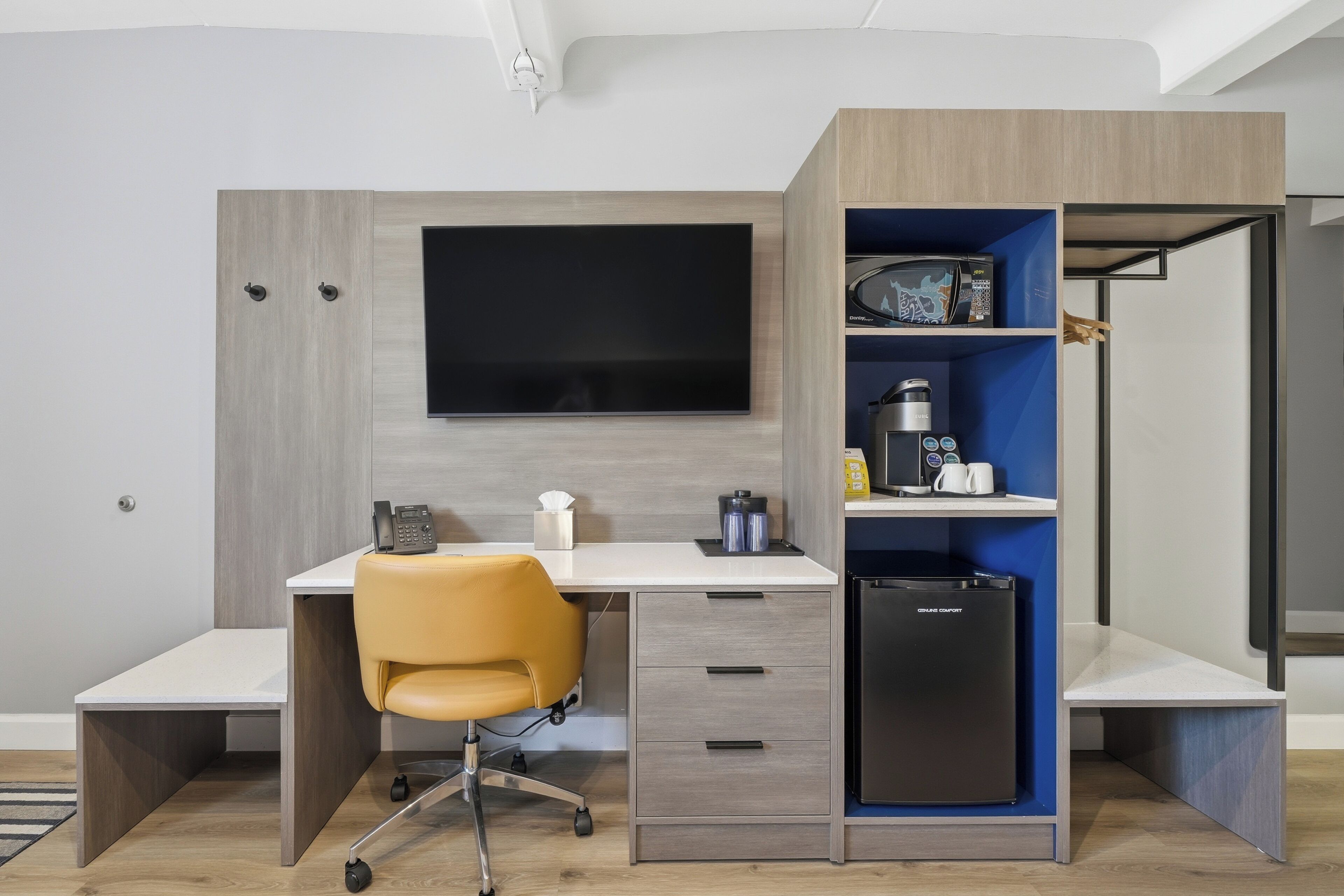 A modern office space with a desk, chair, built-in shelving, a flat-screen TV, and a compact refrigerator.