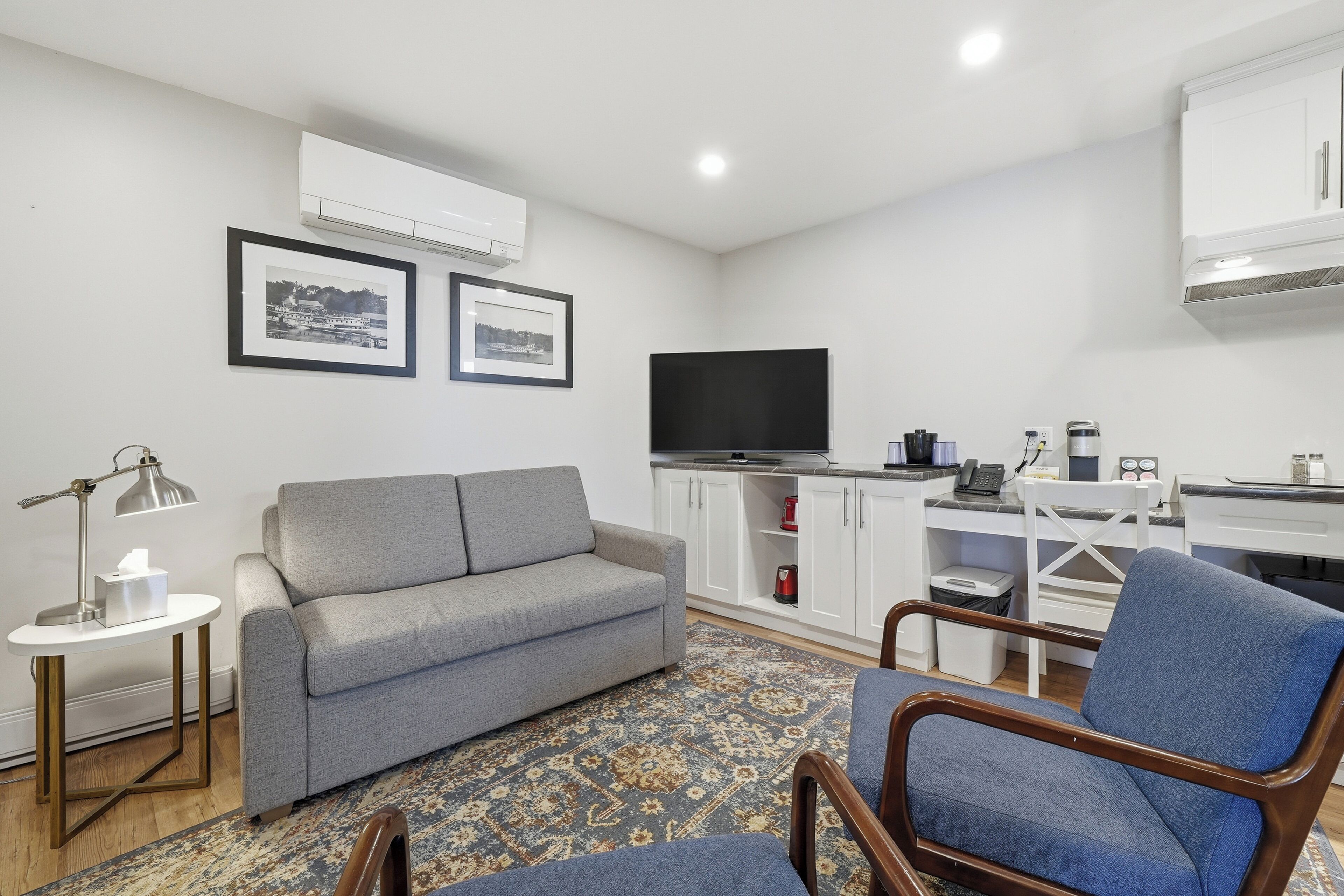 A modern living room with a grey sofa, a blue armchair, a small round table with a lamp, a flat-screen TV, and a kitchen area with white cabinets and a coffee machine.