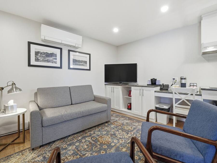 A modern living room with a grey sofa, a blue armchair, a small round table with a lamp, a flat-screen TV, and a kitchen area with white cabinets and a coffee machine.