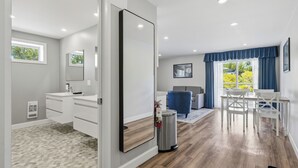 Habitación de hotel moderna con un espejo grande, una cómoda blanca, un cesto de basura pequeño, una zona de comedor con mesa y sillas, y vistas a zonas verdes al exterior.