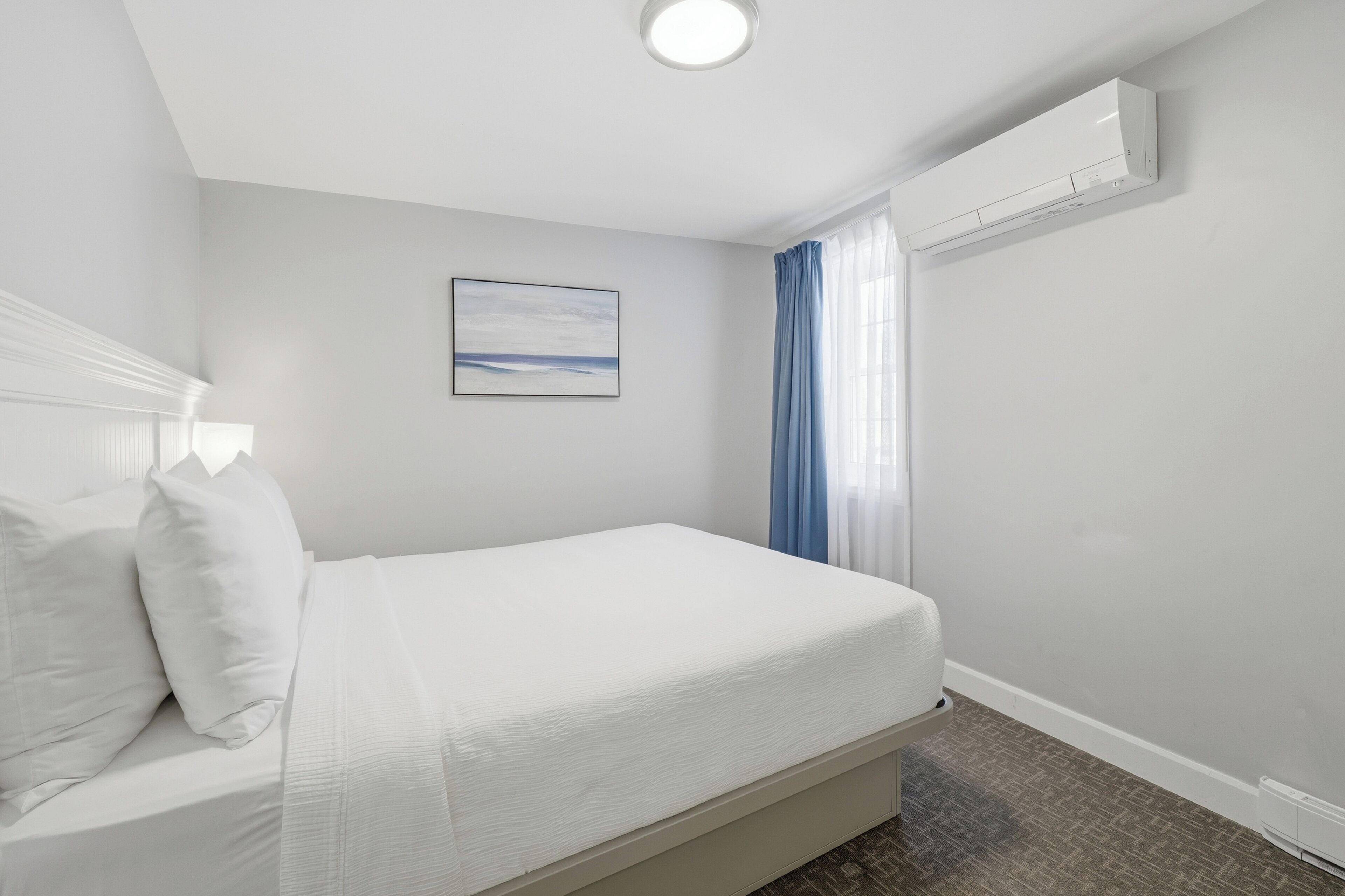 A hotel room with a bed, a ceiling light, an air conditioning unit, and a window with curtains.