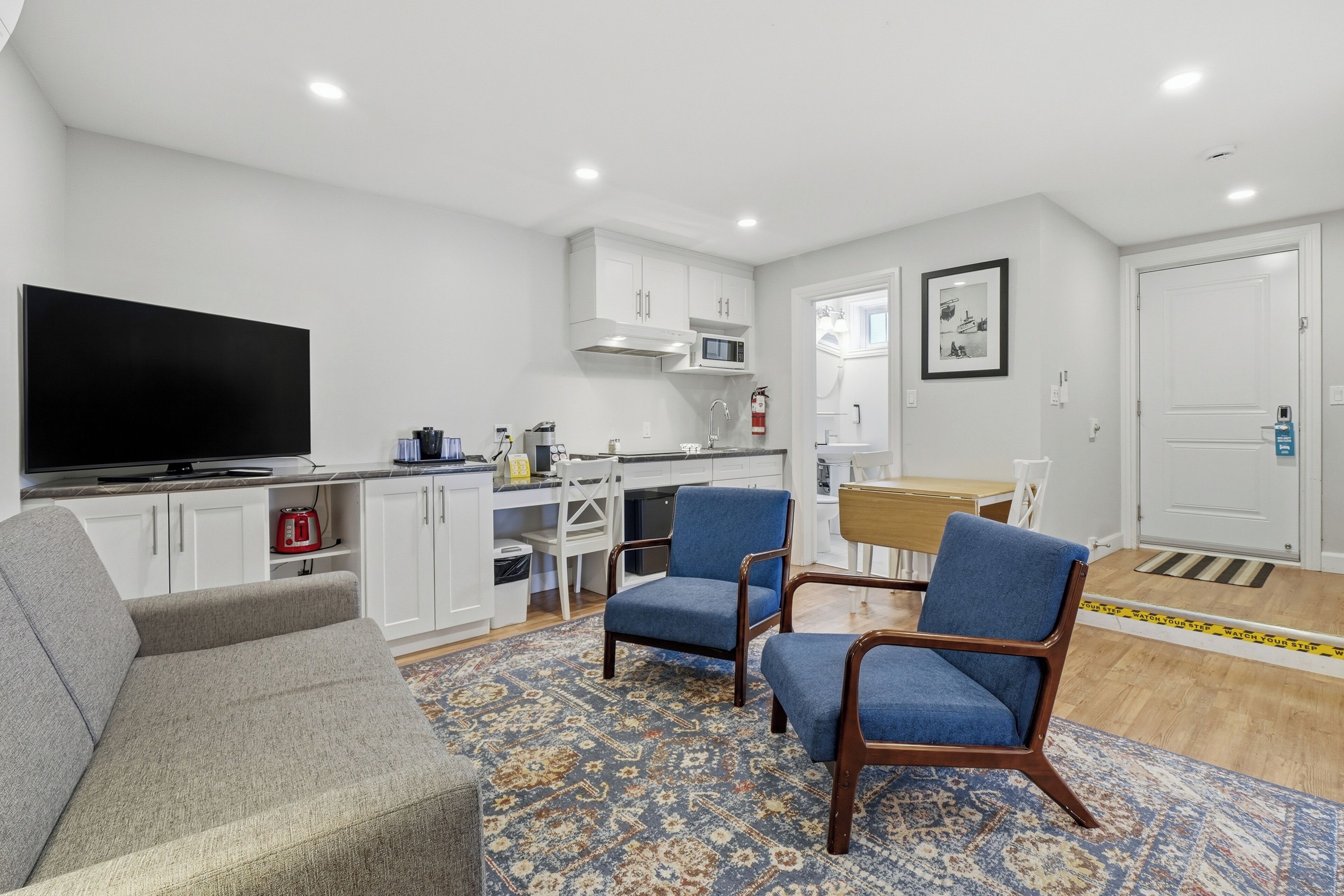 Un salon moderne avec un téléviseur à écran plat, un canapé gris, des fauteuils bleus et un tapis à motifs.