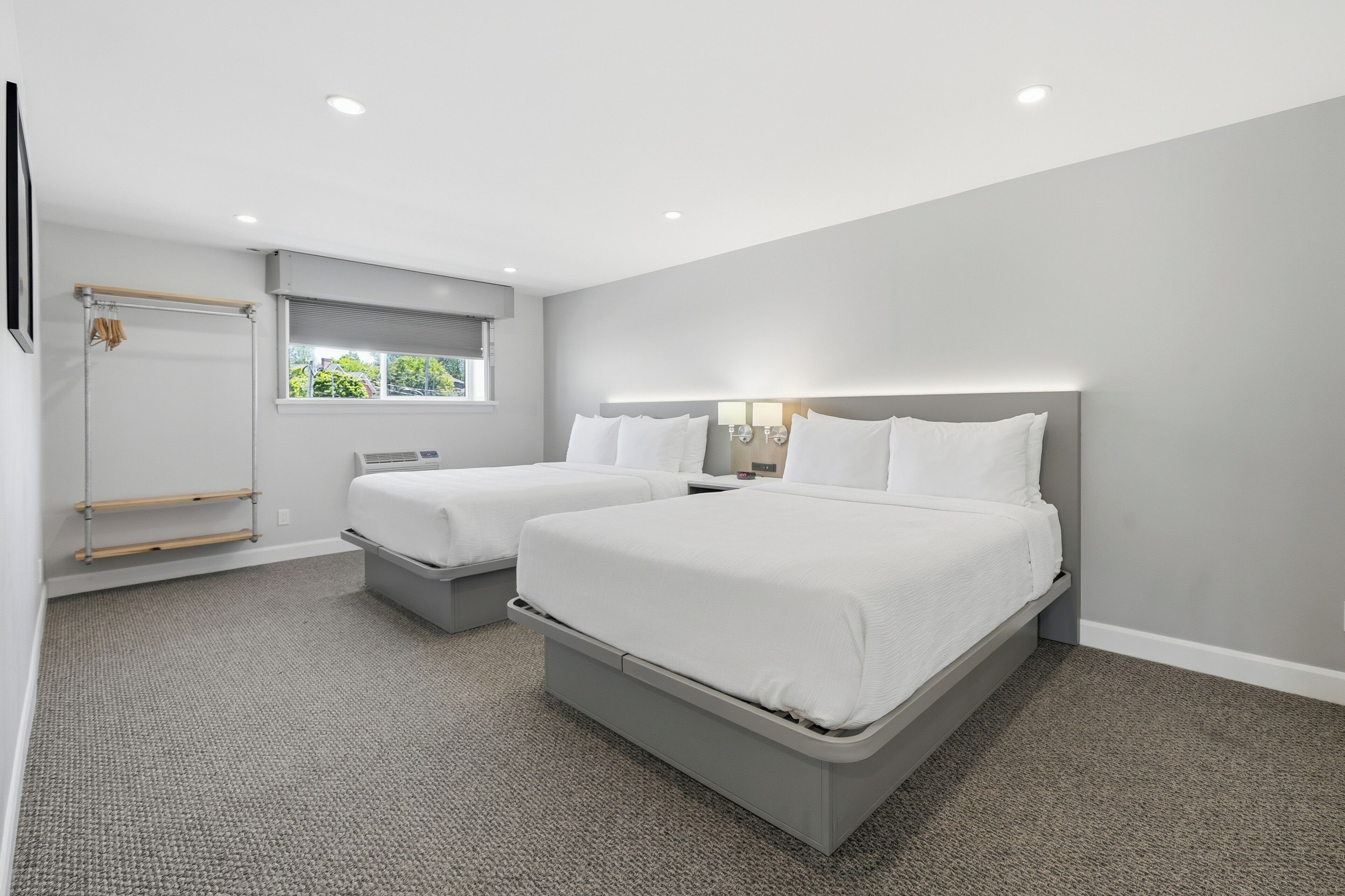 Une chambre d’hôtel moderne avec deux lits, une fenêtre avec stores, un porte-vêtements en bois et un sol recouvert de moquette.