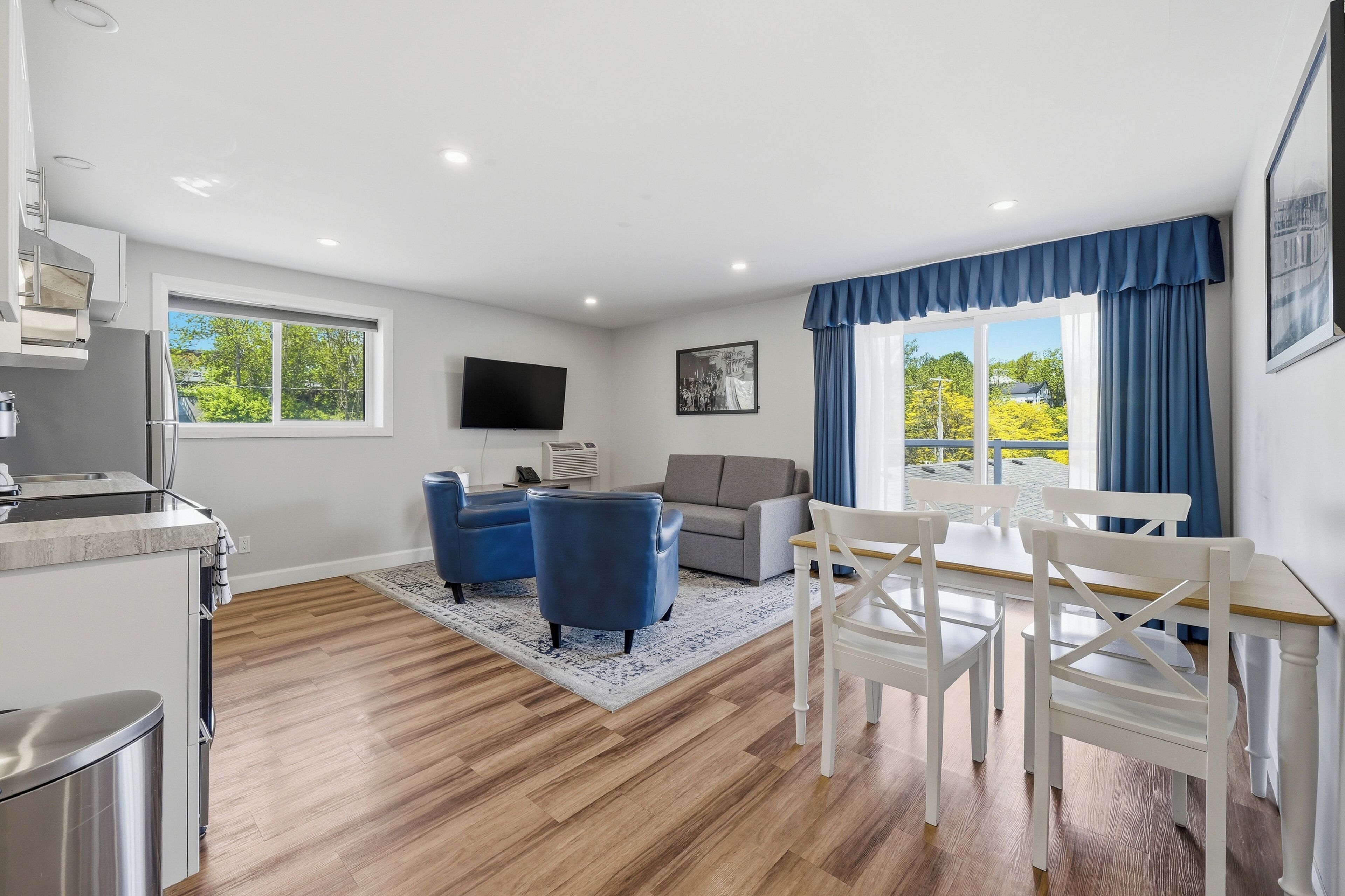 Een moderne woonkamer met een bank, twee blauwe fauteuils, een eettafel met witte stoelen, een flatscreen televisie en een raam met gordijnen.