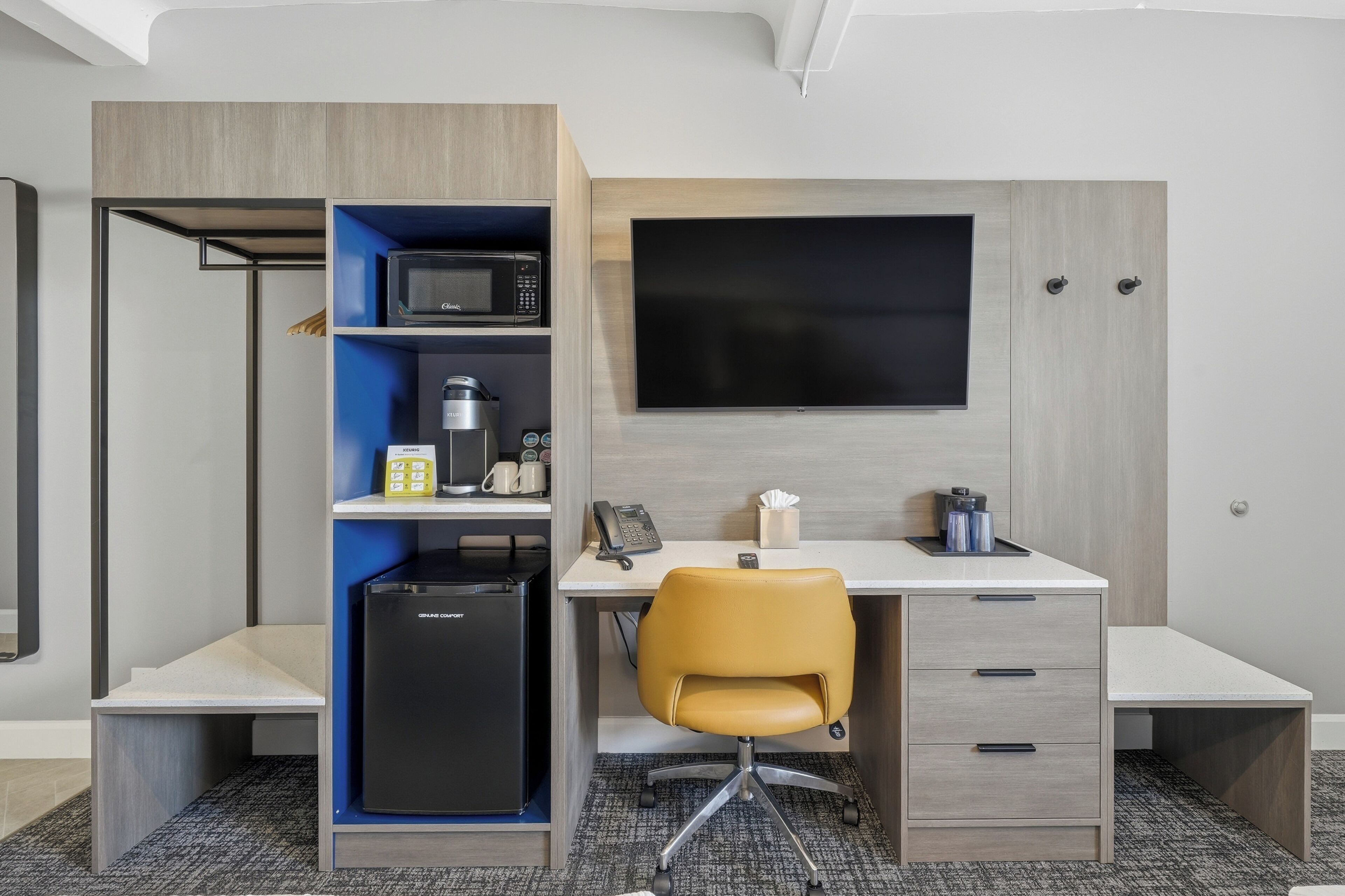 A compact hotel room with a kitchenette, a desk with a chair, a flat-screen TV, and a wardrobe.